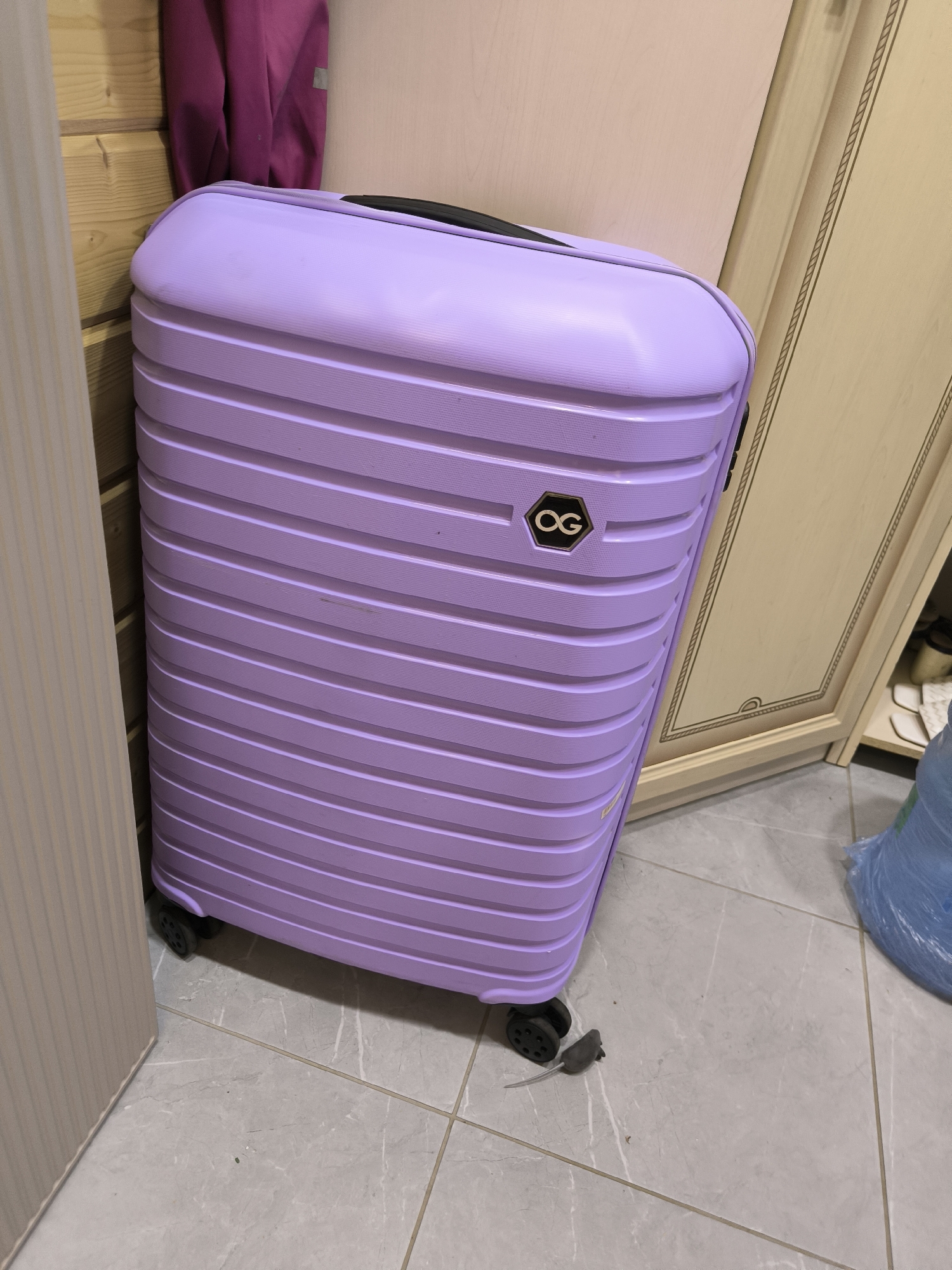 A lavender suitcase stands upright on a light grey tiled floor in a room with light-colored walls. The suitcase has horizontal ribbed detailing and a black handle. A small, dark grey toy mouse is positioned on the floor near the suitcase's wheels. To the left of the suitcase, a wooden wall is visible, and a portion of a dark pink garment hangs on it. To the right, a cream-colored cabinet with decorative trim is present. In the background, a large blue object, possibly a trash bag, is situated on the floor. There are no people in the image. The environment appears to be indoors, possibly a storage area or entryway. There is no information available to confirm the location in the city of Октябрьский, Russia, based on the visual elements alone.