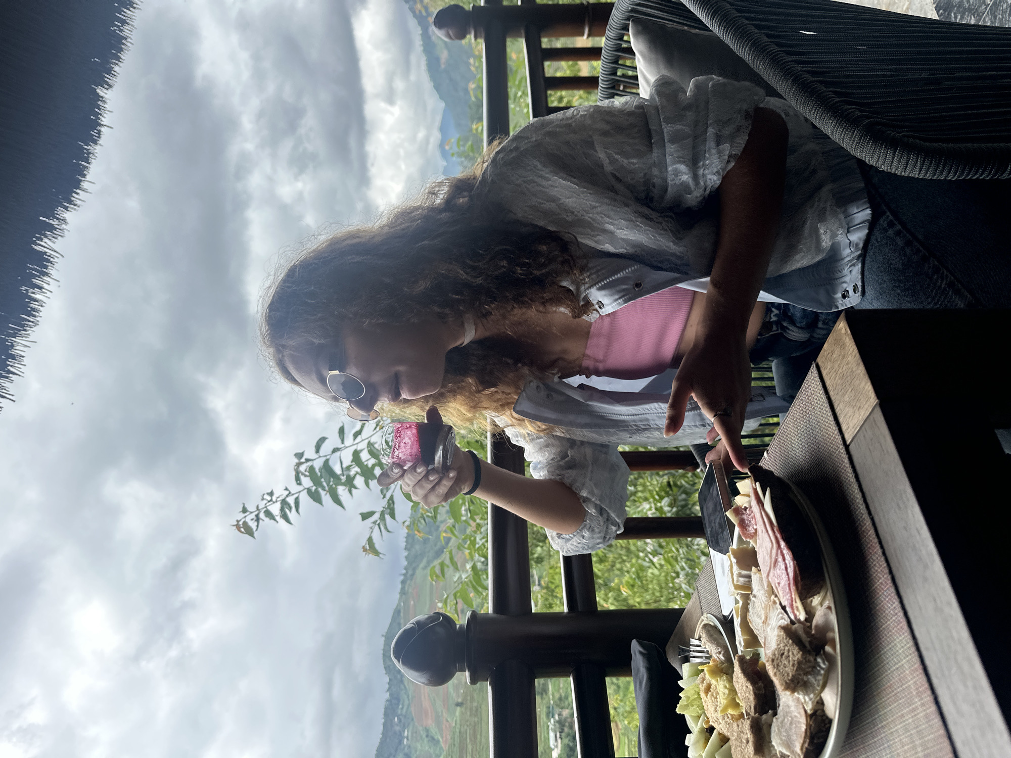 A woman with long, curly brown hair and round, gold-rimmed sunglasses is seated at a dark brown outdoor table on a balcony or terrace in Púng Luông, Vietnam. She is wearing a pink ribbed tube top, a light blue/white sheer long-sleeved button-up shirt worn open, and dark blue jeans. A thin white choker necklace is visible around her neck, and a black elastic band is on her left wrist. She is holding a small, clear glass containing a deep red/purple liquid to her mouth, appearing to drink.
On the table, there is a patterned placemat. A large white oval-shaped plate holds an assortment of open-faced sandwiches with dark bread, various cold cuts (including ham and salami), and soft cheese. A dark-colored smartphone is placed face-down on the table near her right hand. A smaller, partially visible plate to the left contains light green food items (possibly melon or lettuce) and a yellow, scrambled-egg-like dish, with a fork resting on it.
Behind the woman, a dark grey/black woven outdoor chair with a light grey cushion is visible. A dark brown wooden railing with decorative posts separates the dining area from a panoramic view of mountainous terrain, characterized by terraced hills, indicative of agricultural use. Lush green foliage and tree branches are visible through and above the railing. The sky is heavily overcast with grey clouds. A small section of a thatched roof is visible at the very top right of the frame.
