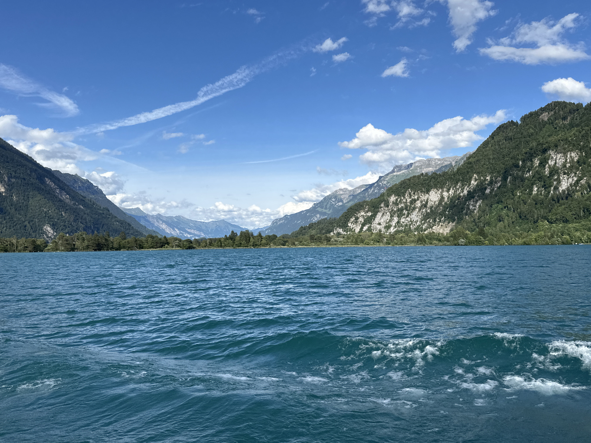 The image shows a wide, turquoise lake with moderate waves in the foreground. In the background, a lush green tree line runs along the shore, backed by steep, rocky mountains. The sky is a bright blue with scattered white clouds and contrails. The scene is serene and natural, with no visible people or man-made structures other than what might be implied by the presence of a boat creating the wake in the water. The overall impression is one of a beautiful, mountainous landscape.