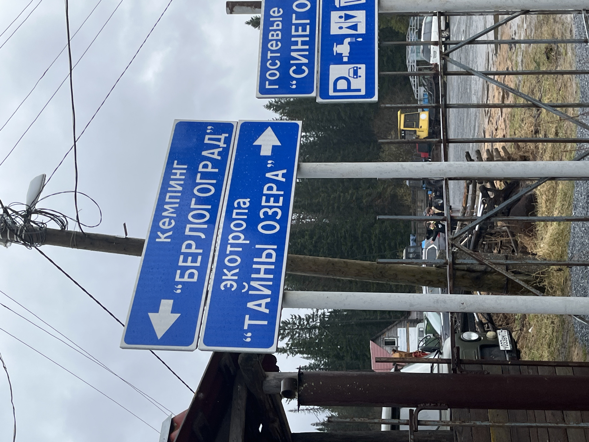 The media file presents an outdoor scene in Zyuratkul, Russia, dominated by several blue informational road signs written in Russian. The image is rotated 90 degrees clockwise. A wooden utility pole with numerous tangled wires is on the left.
The most prominent signs, mounted on metal posts, provide directions. The top-left sign reads "кемпинг БЕРЛОГОГРАД" (camping BERLOGOGRAD) with an arrow pointing towards the bottom of the frame. Below it, a sign for "экотропа ТАЙНЫ ОЗЕРА" (ecotrail SECRETS OF THE LAKE) features an arrow pointing towards the top of the frame. To the right, another blue sign reads "гостевые СИНЕГО" (guest houses BLUE) with an arrow pointing to the left of the frame. Directly below this, a smaller blue sign displays white pictograms for parking (P), a bus or camper van, and a water tap, indicating available facilities.
Behind the signs, a metal fence or gate runs horizontally across the midground. Beyond the fence, various vehicles are visible: a yellow excavator or loader, a white van, and an olive green SUV (possibly a UAZ) with a Russian license plate "0338м 775". Distant buildings and dark coniferous trees are seen in the background. The ground appears to be a mix of dirt, gravel, and sparse grass. The sky is overcast and cloudy, suggesting a dull daytime. The scene indicates a recreational or tourist area, likely near a natural attraction given the mentions of "camping," "ecotrail," and "lake."