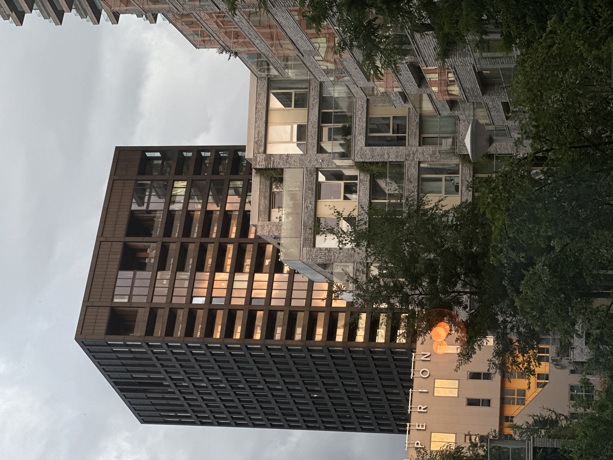 The image depicts a modern architectural complex in Amsterdam, Netherlands, with a prominent dark-colored, multi-story building dominating the left side of the frame. This building features a grid-like pattern of windows and balconies. To its right, a lighter-colored, stepped building with numerous windows and glass balconies is visible, reflecting the warm glow of the setting sun. Further to the right, another building with a brick facade and glass balconies is partially seen. In the foreground, lush green trees obscure the lower portions of the buildings. The sky is overcast with clouds. The building on the far left has the word "PERION" visible in large letters on its facade.