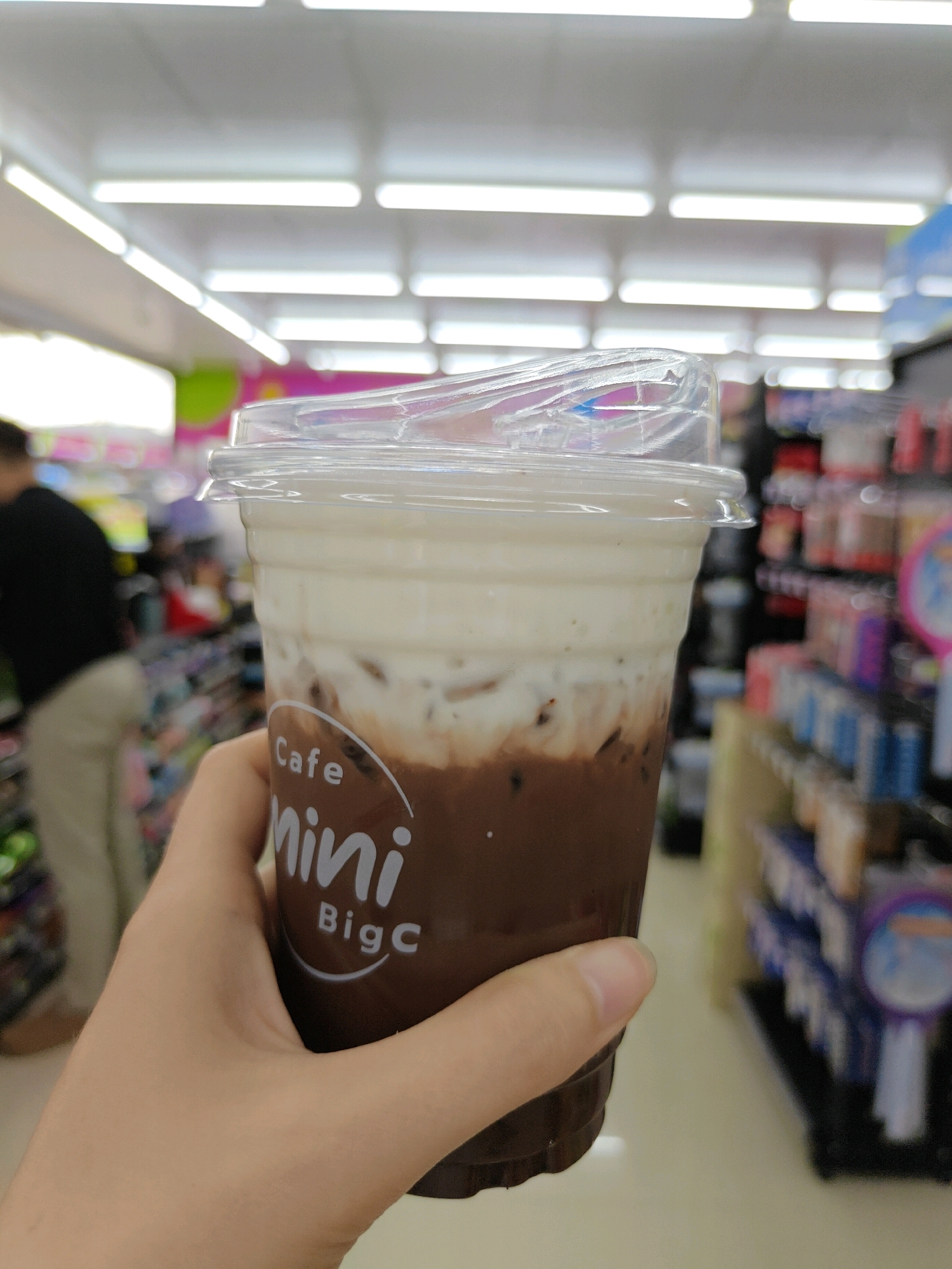 A person's hand holds a clear plastic cup filled with a layered beverage, likely a coffee drink with cream and chocolate. The cup is branded with "Cafe mini BigC" in white lettering. The background shows an aisle in what appears to be a convenience store or supermarket in Mu Si, Thailand, with shelves stocked with products, fluorescent lighting, and a blurred figure of another person in the distance. The scene is brightly lit and suggests an indoor retail environment.