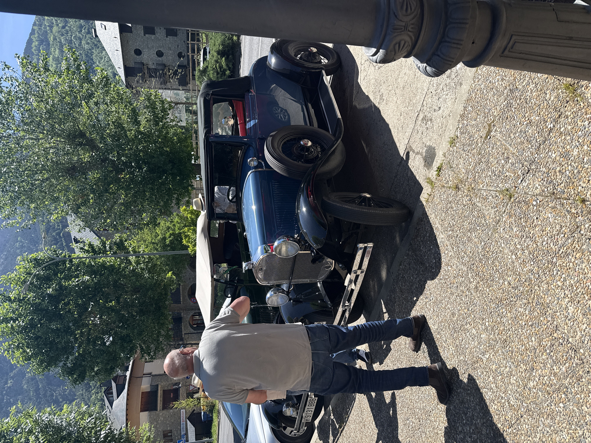 A man in a gray t-shirt, blue jeans, and brown shoes stands next to a dark blue vintage car, possibly a Model A Ford, with a spare tire mounted on the side. A white car is parked in front. The man appears to be looking at the front of the vintage car. The background includes trees, a stone building with arched windows, and mountains. The ground is paved with small stones or pebbles. The photo appears to be taken on a sunny day. A post, likely part of a fence or street feature, is visible on the right side. The scene suggests it's likely located in the town of Benasque, Spain, given the architecture and mountainous backdrop.