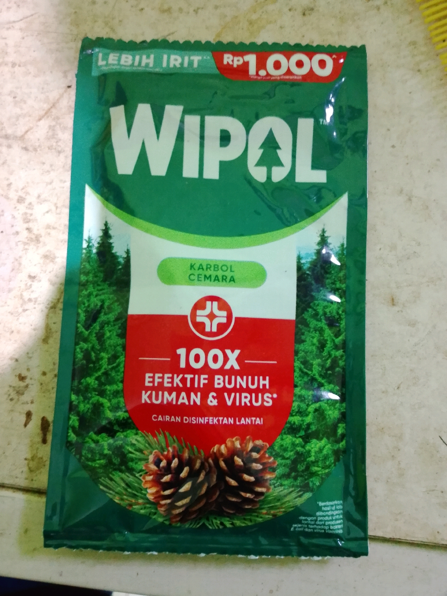 The image shows a close-up of a green, foil-like sachet of "WIPOL" brand disinfectant. The sachet features a pine tree motif, and text in Indonesian. Key text includes "LEBIH IRIT RP 1.000" (more economical RP 1,000), "WIPOL" in large white letters with a stylized pine tree as the "O", "KARBOL CEMARA" (pine carbolic), and "100X EFEKTIF BUNUH KUMAN & VIRUS" (100X effective to kill germs & viruses). Below this, it states "CAIRAN DISINFEKTAN LANTAI" (floor disinfectant liquid). Two pine cones are depicted at the bottom of the sachet. The sachet is placed on a speckled, light-colored surface, possibly a countertop or floor, with visible texture. The lighting suggests an indoor environment, possibly with some natural light contributing to glare on the sachet. There is no visible indication of the city of Gebang, Indonesia, within the image itself.