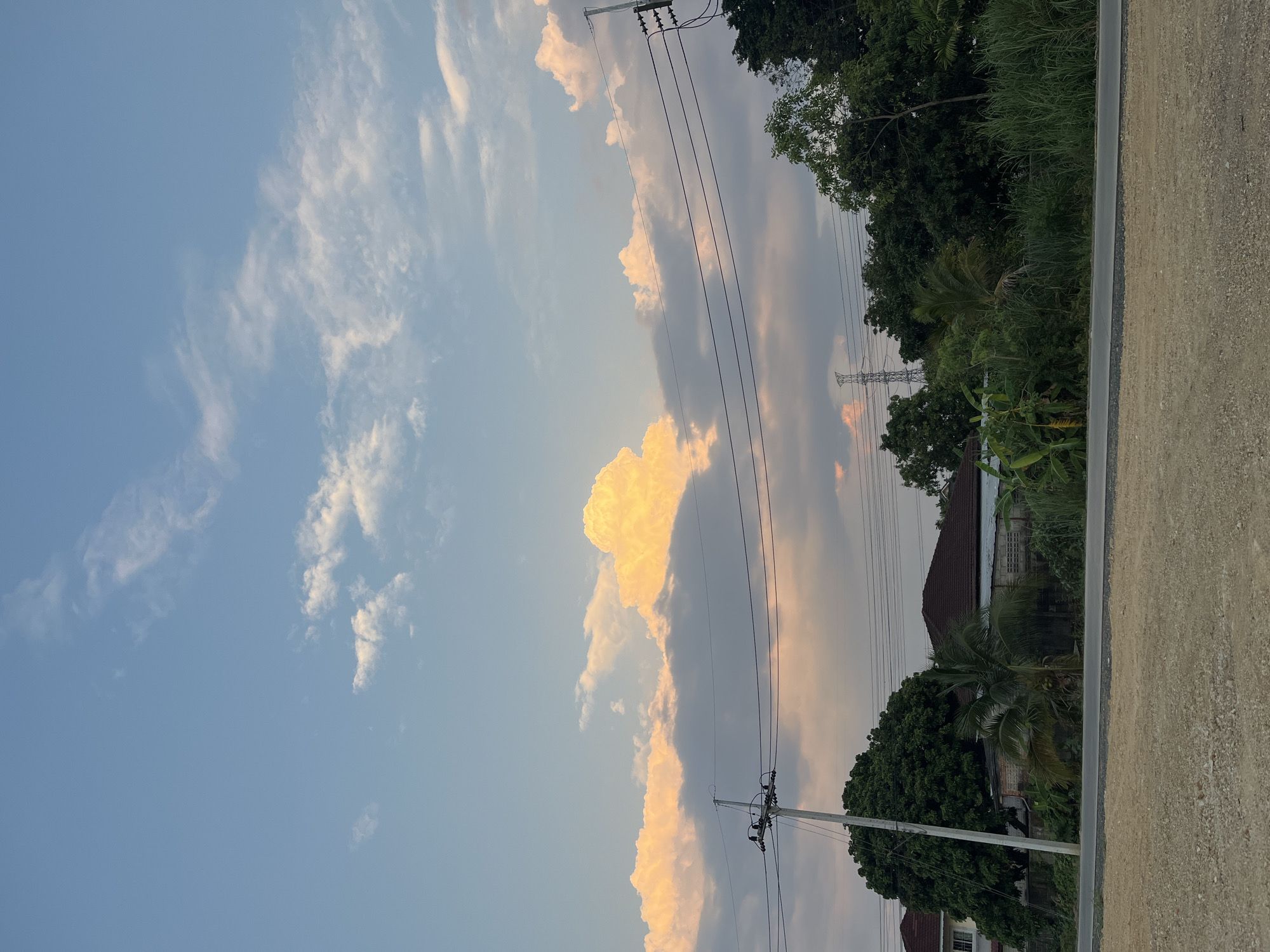 The image shows a daytime sky with clouds illuminated by the sun, likely during sunset or sunrise. The location appears to be in a suburban or rural area of บ้านกาญจน์กนก 23, Thailand. In the background, there are houses and lush green vegetation, including trees and bushes. Overhead power lines run across the frame. A portion of a paved sidewalk or road is visible on the right edge of the photo. No people are present in the image.