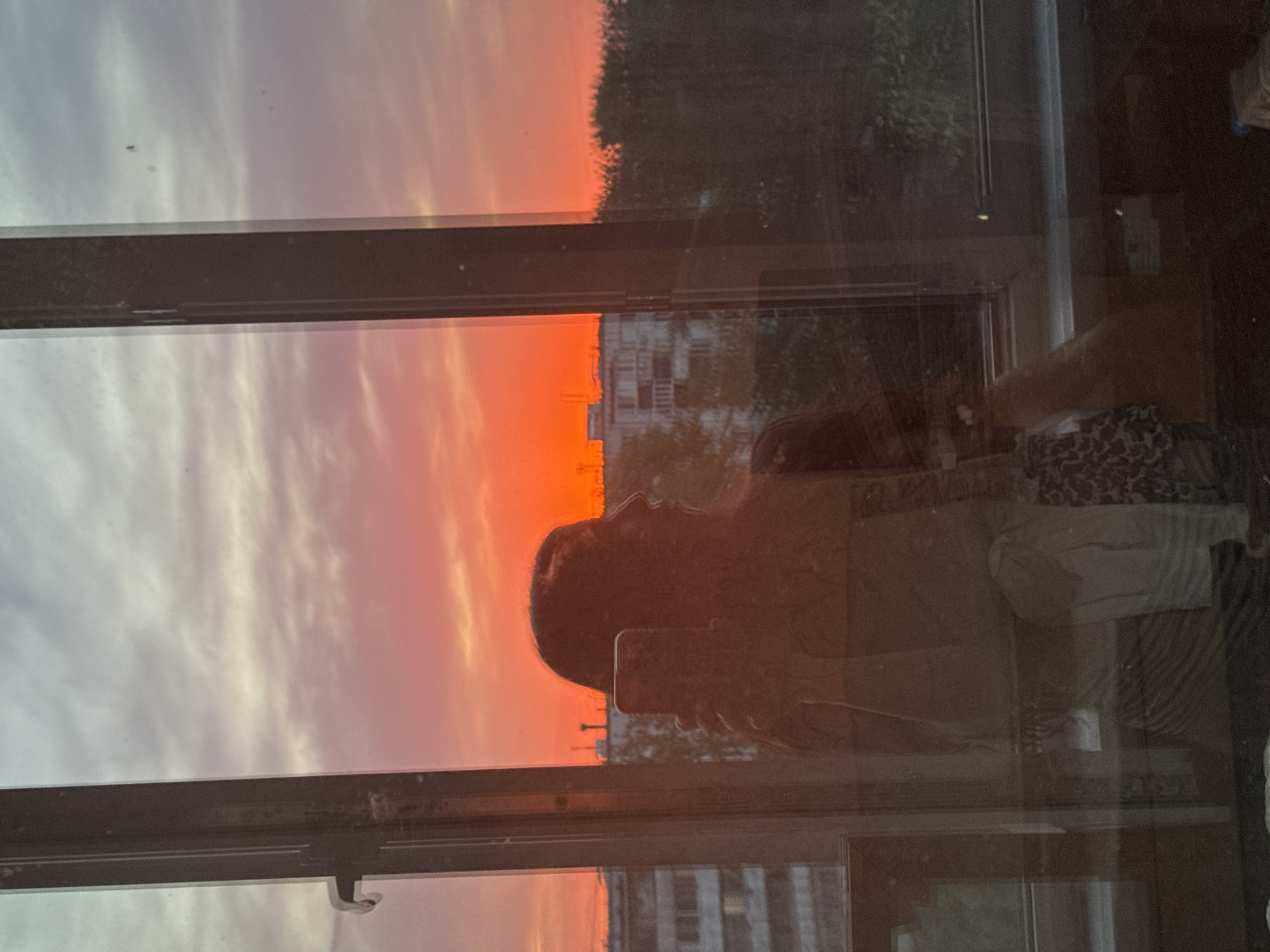 The image shows a sunset reflected in a window. Outside the window, buildings are visible. The reflection includes the person taking the picture, holding a phone. The buildings suggest a relatively urban environment. Without further information, a specific location in Kazan, Russia, cannot be identified.
