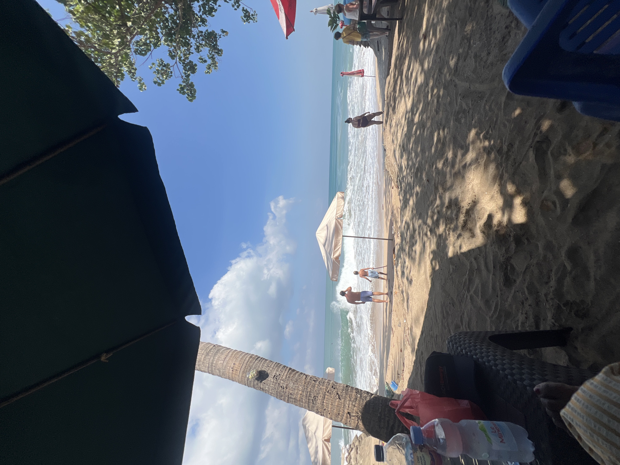 The image captures a sunny day at Kuta Beach, Indonesia, from a shaded viewpoint under a large dark umbrella. In the foreground, a person's feet covered with a striped towel are visible, along with a black patterned chair and a water bottle labeled "Le Mineral." A red bag and a black baseball cap are also on the chair.
The beach itself is sandy, with waves crashing onto the shore. Several beach umbrellas are scattered across the sand. In the middle ground, a large palm tree stands to the left, and two men are looking out at the ocean. One man, wearing blue swim trunks, stands closer to the water, while a younger boy in a light blue shirt and shorts stands further back with a stick.
Further down the beach, more people are visible. A woman in a dark bikini stands facing the ocean. To the right, a few individuals are seated at tables under a red and white umbrella, with some sitting at a wooden bench. The ocean is a vibrant blue, with whitecaps from the waves. The sky is clear blue with scattered white clouds. Lush green trees are visible on the right side of the image, bordering the beach. The overall scene depicts a lively day at a popular tourist beach.
