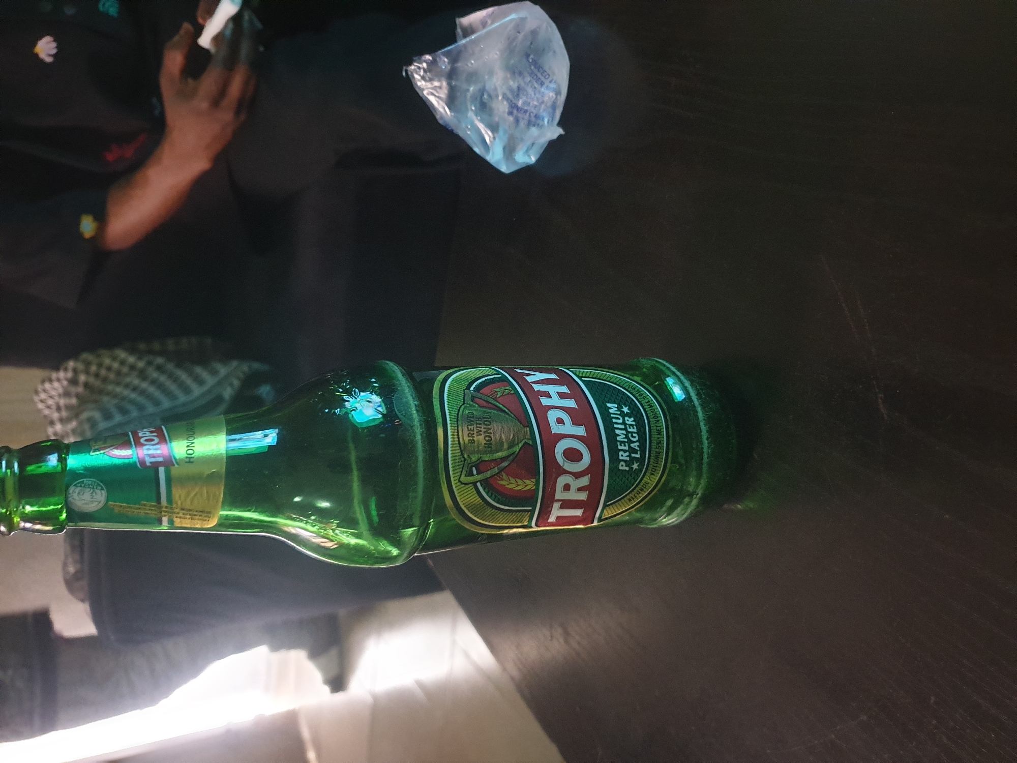 A green glass bottle of Trophy Premium Lager beer stands upright on a dark wooden surface. The bottle's label is visible, featuring a trophy graphic and the words "Trophy Premium Lager." In the background, a person is seated on a couch, holding a white object, possibly a cigarette or a tissue. The environment appears to be indoors, with soft lighting casting shadows. The scene is undated and location is unknown.