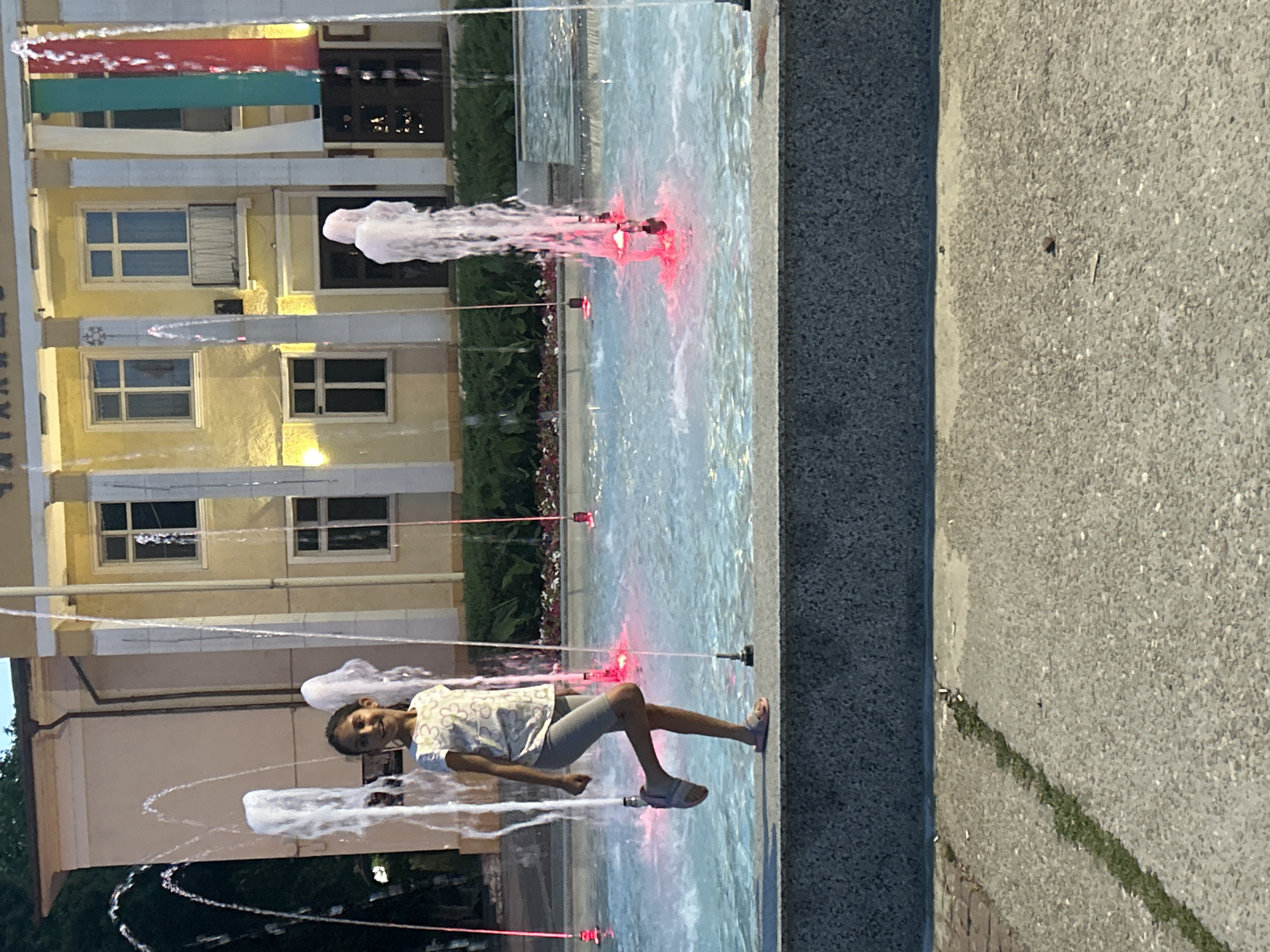 A young girl with dark hair, wearing a white t-shirt with a floral pattern and grey shorts, is standing in a fountain. She is looking towards the camera and smiling, with her left leg bent at the knee and her right foot resting on the fountain's edge. The fountain has multiple water jets, some illuminated with red lights, creating a dynamic visual. Behind the fountain, a building with a light-colored facade and several windows is visible. Vertical banners in white, green, and red hang from the building, suggesting a national or regional affiliation. The environment appears to be an outdoor public space, likely a town square or plaza, in Parvomay, Bulgaria. The ground in front of the fountain is paved with textured material, with a strip of grass running through it.