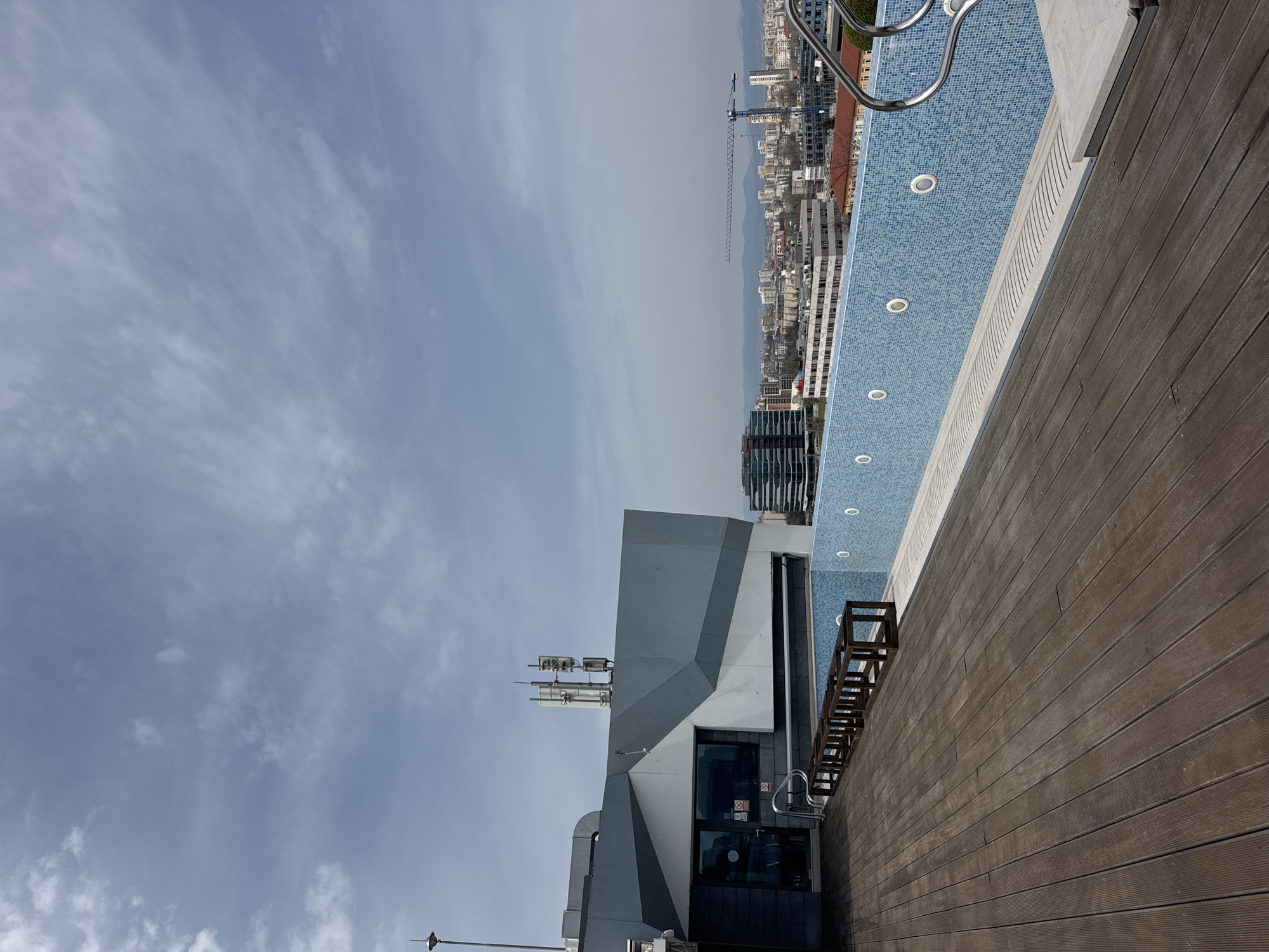 The image shows a high-angle view from a building, likely a rooftop in the city of Sofia, Bulgaria. The sky is overcast with light-colored clouds. A section of a rooftop pool is visible, with blue tiles and recessed lights. A wooden deck surrounds the pool. The cityscape is visible in the distance, including buildings, a construction crane, and mountains. A building with a unique architectural design featuring antennas is in the foreground.