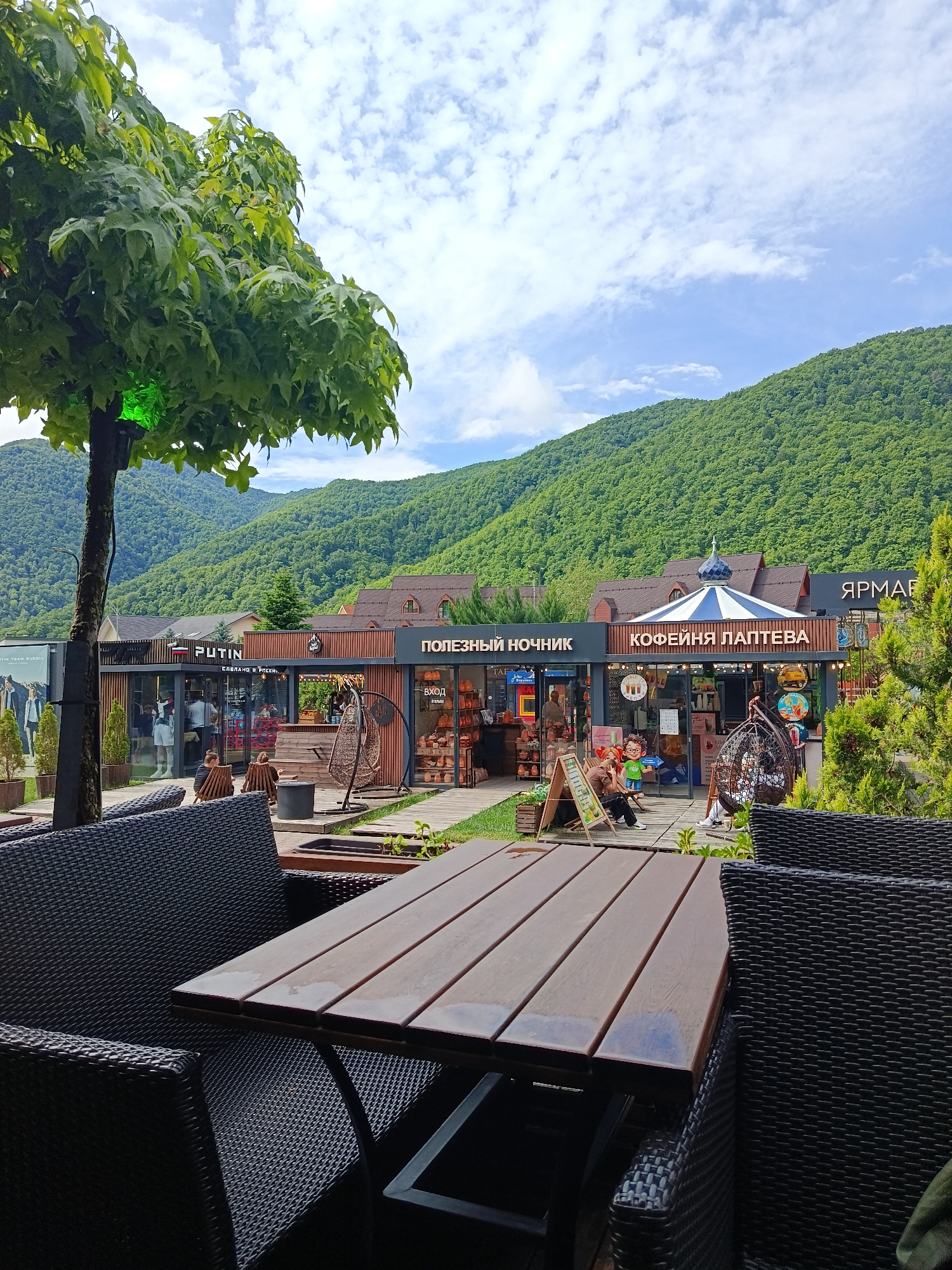The image shows an outdoor seating area in front of shops with a mountainous backdrop. The seating area features dark wicker chairs and a wooden table in the foreground. In the middle ground, there are several small shops with signage in Russian. One shop has a sign that reads "PUTIN". Another shop is a cafe named "КОФЕЙНЯ ЛАПТЕВА" (Lapteva Cafe). There are people browsing and sitting in the shops and surrounding areas. A large tree with green foliage is to the left of the frame. The background consists of lush green mountains under a partly cloudy sky. The location is identified as Estosadok, Russia.