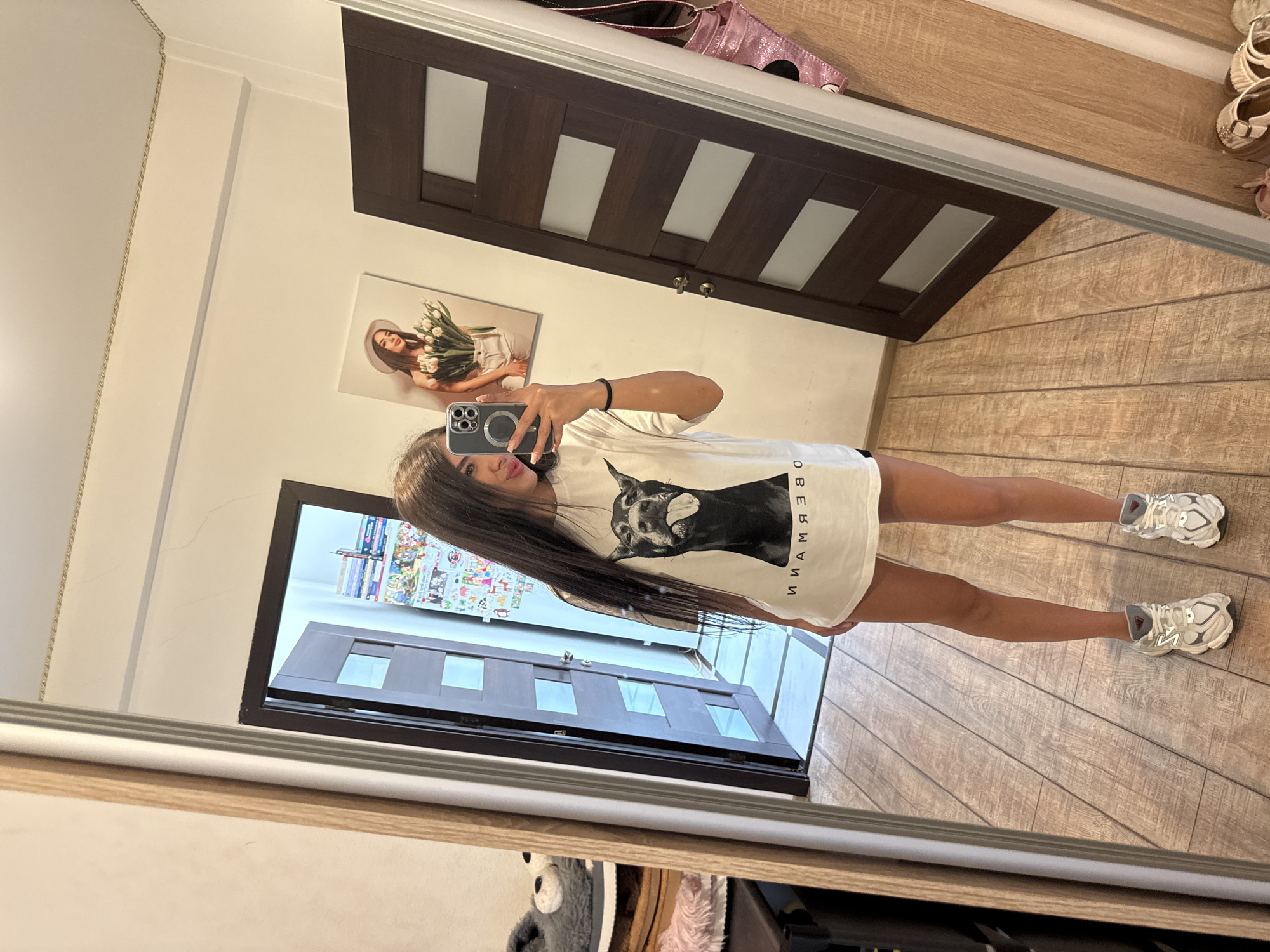 A young woman with long, dark hair is taking a selfie in a mirror. She is wearing a white t-shirt with a black dog graphic and text, black shorts, and white sneakers. She is holding a smartphone in her right hand, which is angled to capture her reflection. In the reflection, we can see a doorway with a dark wood door with frosted glass panels, and a painting of a woman holding flowers on the wall. To the left of the woman, there is a large mirror, and behind her, there are shelves with various items. The floor is made of wooden planks. The scene appears to be an indoor setting, possibly a home. There is no specific information in the image to indicate it is located in Chișinău, Moldova.