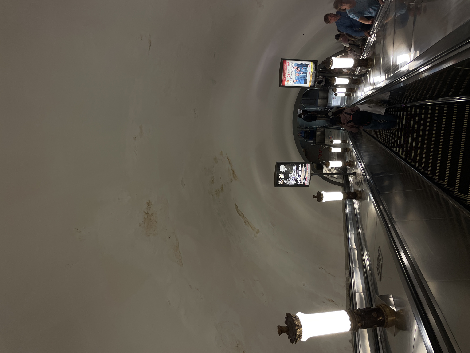 The image depicts an interior scene of a subway station in Saint Petersburg, Russia, specifically an escalator going down. The ceiling shows signs of water damage with discoloration and peeling paint in several areas. On the left side, a large ornate lamp with a white illuminated glass shade is visible, and further down, another similar lamp. On the right side, at the top of the escalator, are two digital advertising screens. The lower screen displays a poster for "IMPROVIZATORS" with an age restriction of 18+. Several people are seen on the escalators, some going down and some going up. The escalators themselves are wide and made of polished metal with black ribbed steps. The overall atmosphere is dimly lit, with the primary light sources being the escalator lights and the advertising screens.