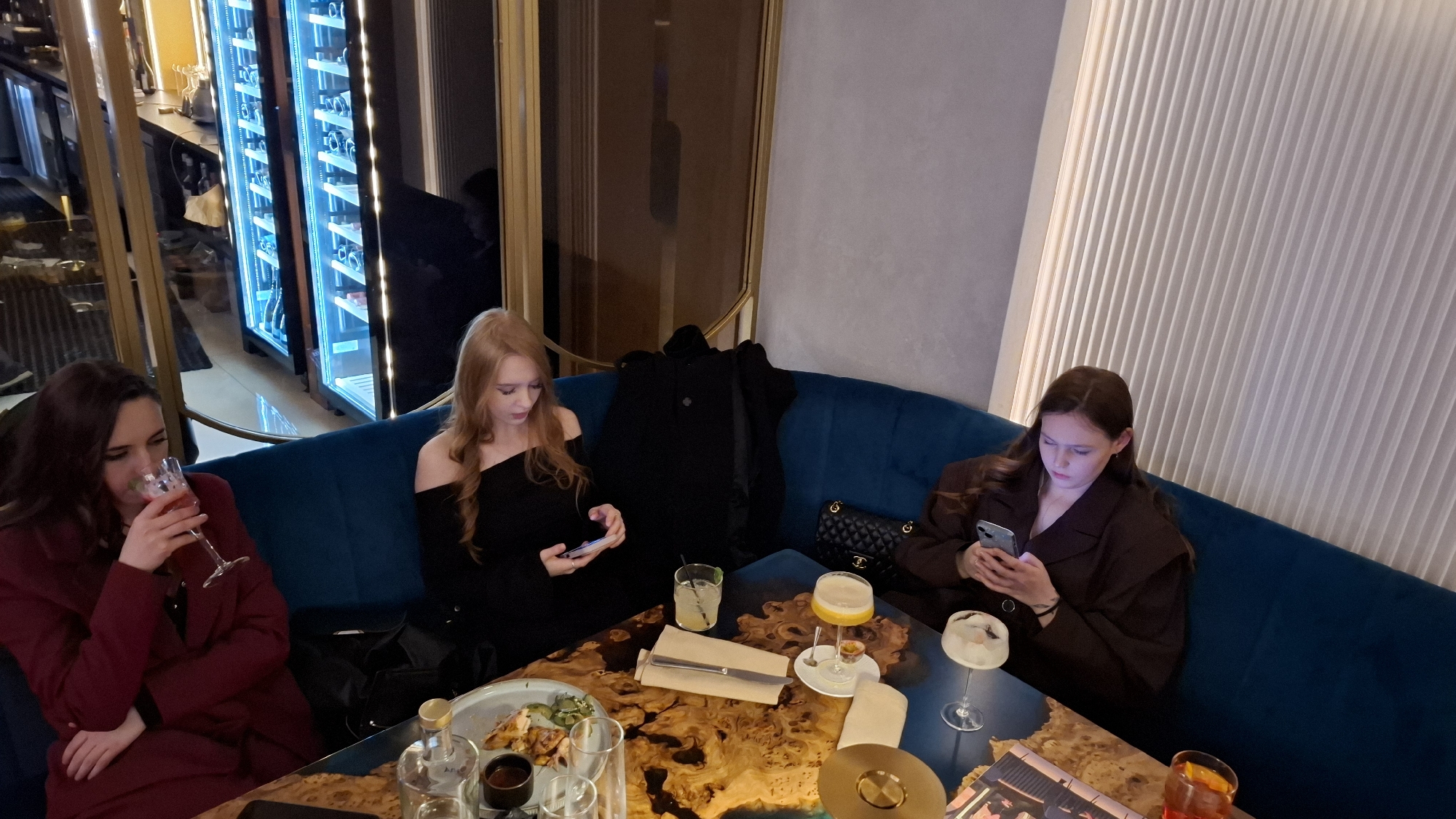 Three women are seated at a table in a restaurant. The woman on the left is wearing a maroon coat and drinking from a stemmed glass. The woman in the center is wearing a black off-the-shoulder dress and looking at her phone. The woman on the right is wearing a brown blazer and also looking at her phone. The table has a unique swirling pattern and several drinks and dishes are arranged on it. A black coat is draped over the back of the booth between the two women in the center and right. A black quilted purse is also visible next to the woman on the right. The booth seating is deep blue velvet. The background consists of a ribbed wall on the right, and a lit display case containing bottles on the left. A mirror is also visible. The location is Saint Petersburg, Russia.
