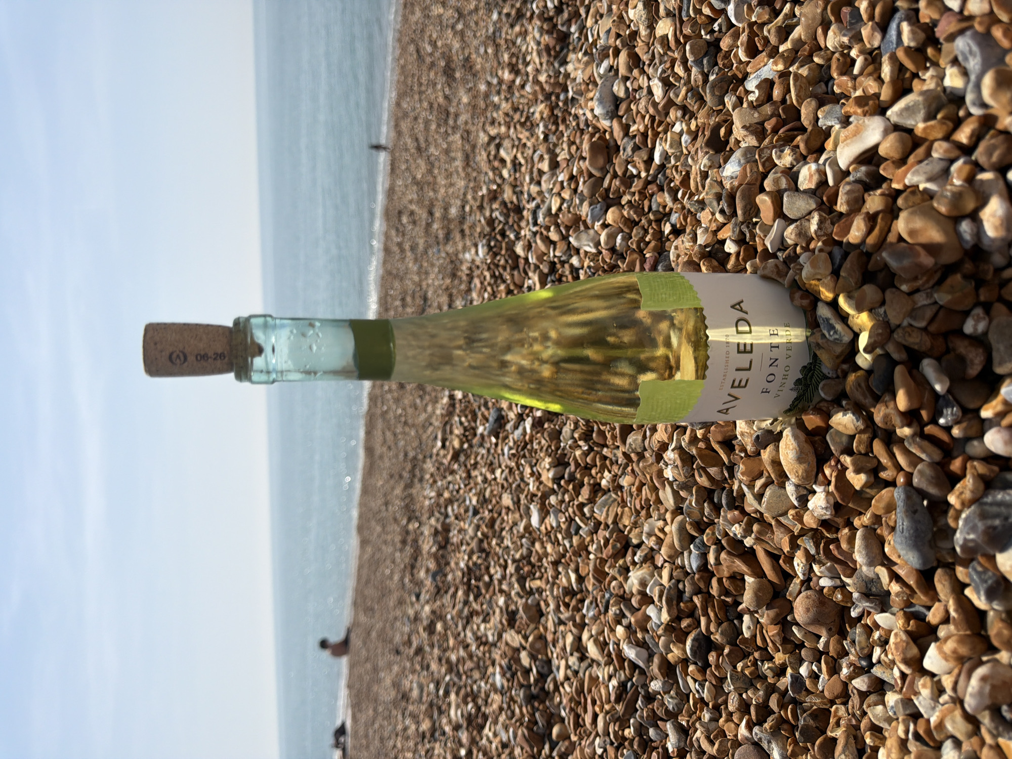 The image shows a bottle of Avelada Vinho Verde wine lying on a pebble beach. The bottle is oriented horizontally with the cork facing left and the label facing up. In the background, the sea and sky are visible with a few indistinct figures on the beach near the water. The pebbles are predominantly brown and grey, varying in size and shape. The location is likely a beach within the Folkestone and Hythe District, United Kingdom.