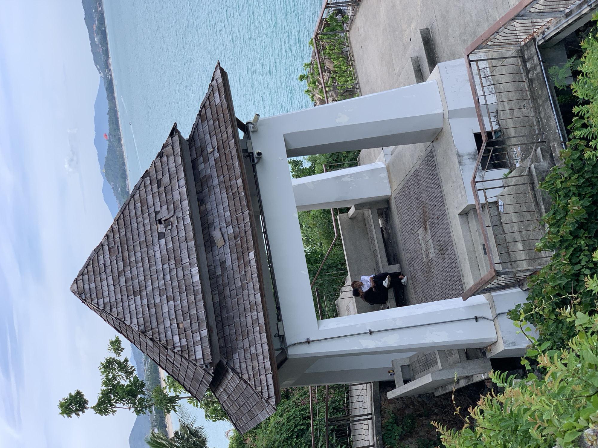 The image depicts an open-air pavilion with a weathered wooden shingle roof, situated on a hillside overlooking a turquoise ocean. Two individuals, one wearing a dark shirt and the other a white shirt, are seated closely together on a stone bench within the pavilion. The pavilion has white concrete pillars and a grey concrete floor, with metal railings along the edges of the surrounding paved area and stairs. Lush green vegetation borders the scene, with palm trees visible in the distance. The ocean extends to a horizon marked by rolling hills and a cloudy sky. A parasail can be seen in the sky over the water. This scene is likely located in Baan Chaweng Noi, Thailand, known for its coastal beauty and recreational activities.