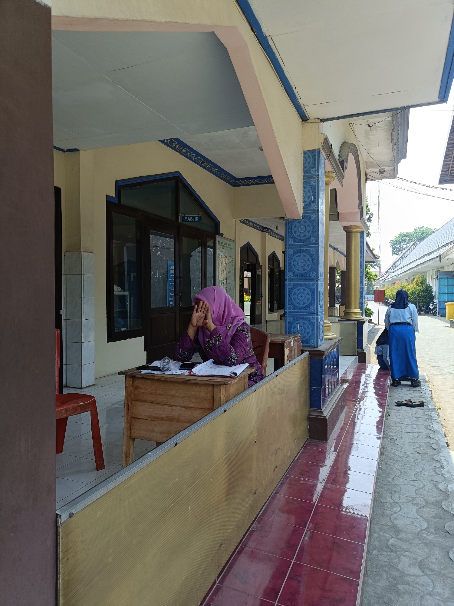 The image depicts an outdoor scene, likely in Kediri City, Indonesia, characterized by the architectural style of the building and the presence of individuals dressed in modest attire. In the foreground, a person wearing a purple hijab and matching tunic is seated at a wooden desk, with their hands covering their face, suggesting an emotional state of distress or exhaustion. The desk is cluttered with papers and a plastic cup. To the right, another individual, also in modest attire with a blue hijab and skirt, stands facing away from the camera, observing something in the distance. The building features a covered walkway with tiled flooring and decorative blue tile accents. A red chair is visible to the left of the desk. The environment appears to be a public space, possibly a school or administrative building, with a clear pathway and a glimpse of greenery in the background.