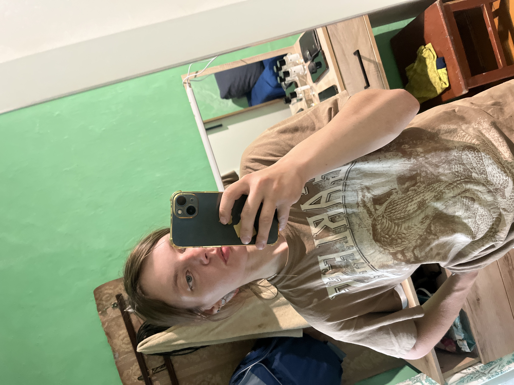 A person is taking a selfie in a room with green walls. The person is holding a dark green smartphone with a gold trim. They are wearing a brown t-shirt with a graphic design. The background includes a mirror, a cabinet with drawers, and various items stored inside the open drawers. The location is a room, likely in a residential building. The exact location of the city of Nizhny Tagil, Russia is not discernable from the image.
