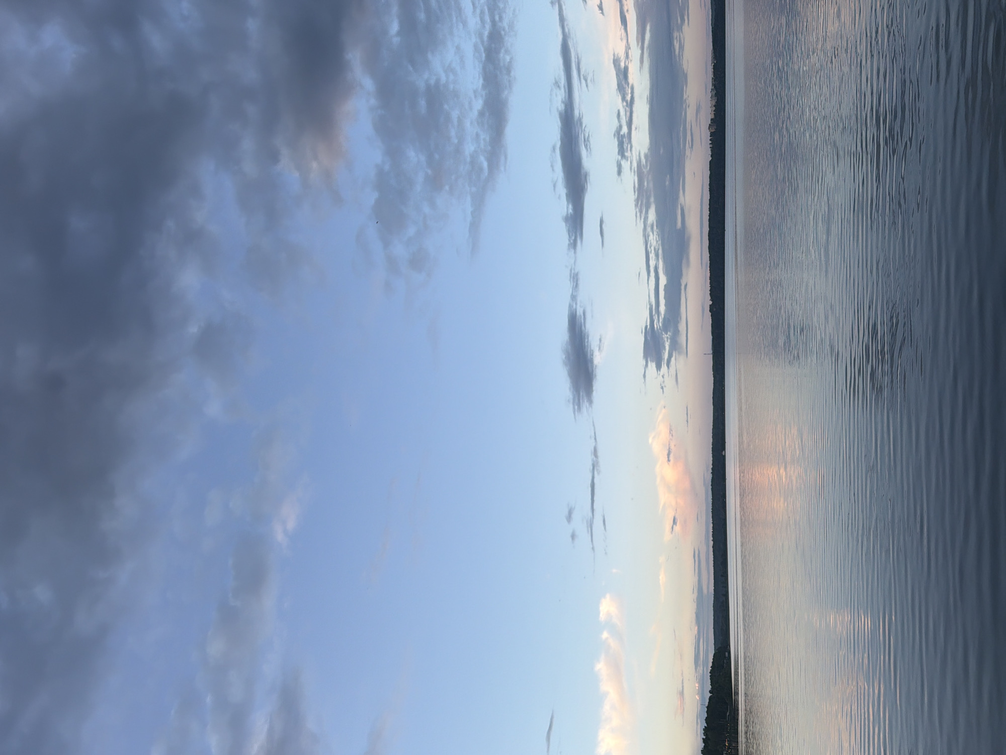 The image depicts a serene body of water, likely a lake or a large river, with a distant tree-lined horizon. The sky is a mix of soft blue and scattered grey clouds, with hints of pink and white suggesting either sunrise or sunset. The water's surface is calm, reflecting the colors of the sky and creating a ripple effect. There are no people or visible structures on the water. The scene is peaceful and natural, characteristic of an outdoor environment near Solnechnogorsk, Russia, potentially overlooking Lake Senezh.