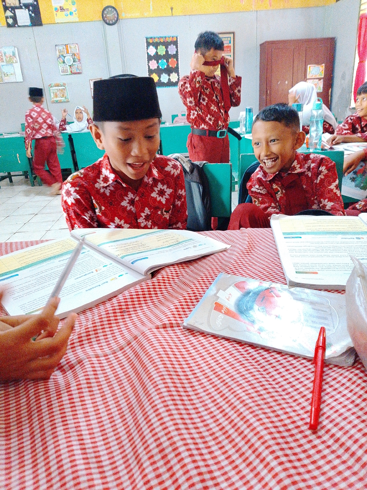 In a classroom setting in Tembung, Indonesia, two young boys in matching red patterned shirts and dark peci hats are the central focus, engaged with books on a red and white checkered tablecloth. The boy on the left, wearing a black peci, is looking at a book open before him, his mouth agape in a smile or laugh. A hand is visible in the lower left corner, holding a pencil-like object over the book. To the right, another boy, also in a red patterned shirt, grins widely, looking slightly upwards. Behind them, another student in a white hijab can be seen, and further back, a boy is adjusting a red tie around his neck. The classroom environment is suggested by green desks, posters on the wall, a clock, and a cabinet. The lighting is somewhat diffused, creating a warm atmosphere.