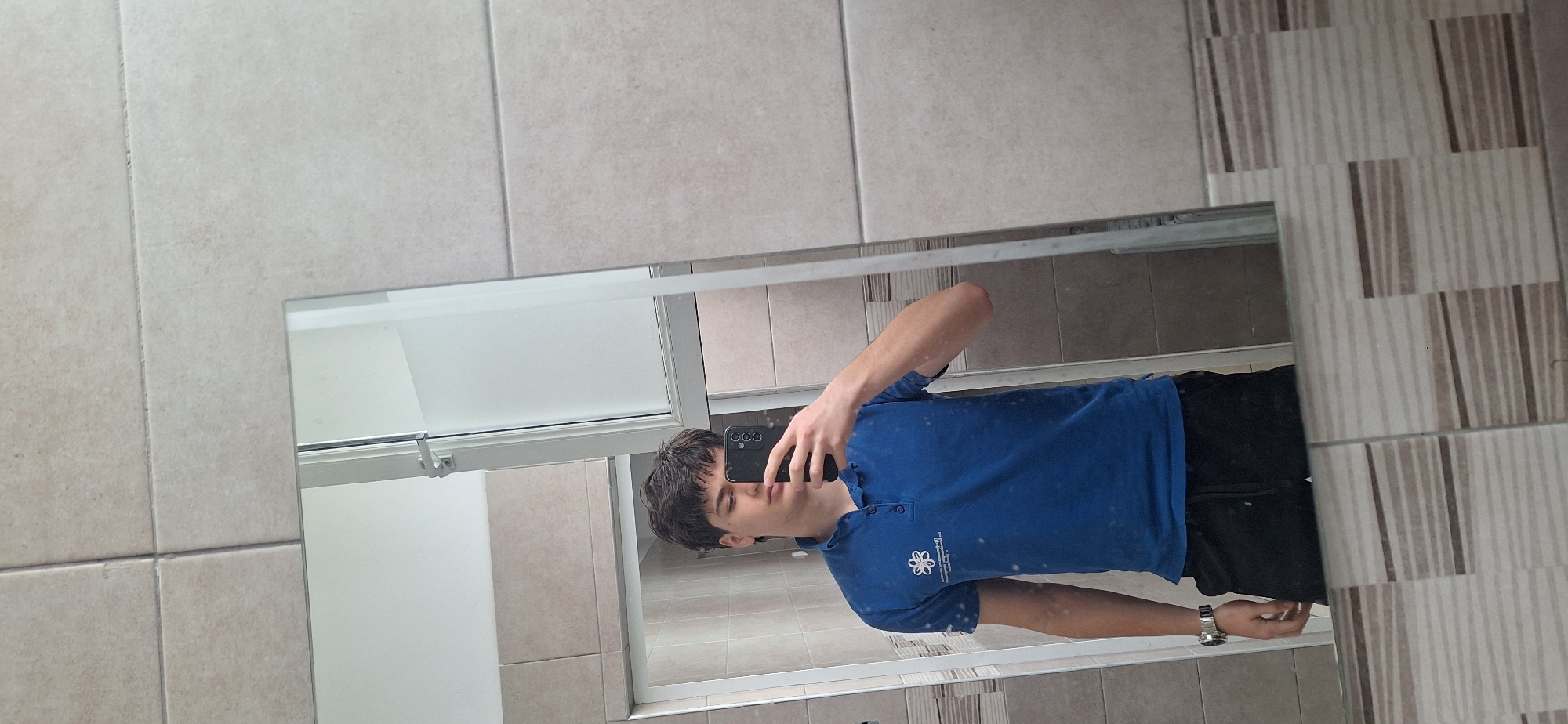 A young male is taking a selfie in a bathroom mirror. He is wearing a blue polo shirt with a logo on the left breast and black shorts. He is holding a black smartphone in his left hand. A watch is visible on his left wrist. The bathroom has light tan tile on the walls behind the mirror and vertical striped tile to the right of the mirror. The mirror reflects the interior of a shower, including a shower door. It is unclear if this photograph was taken in Burgas, Bulgaria. There is no contextual information that indicates the location.