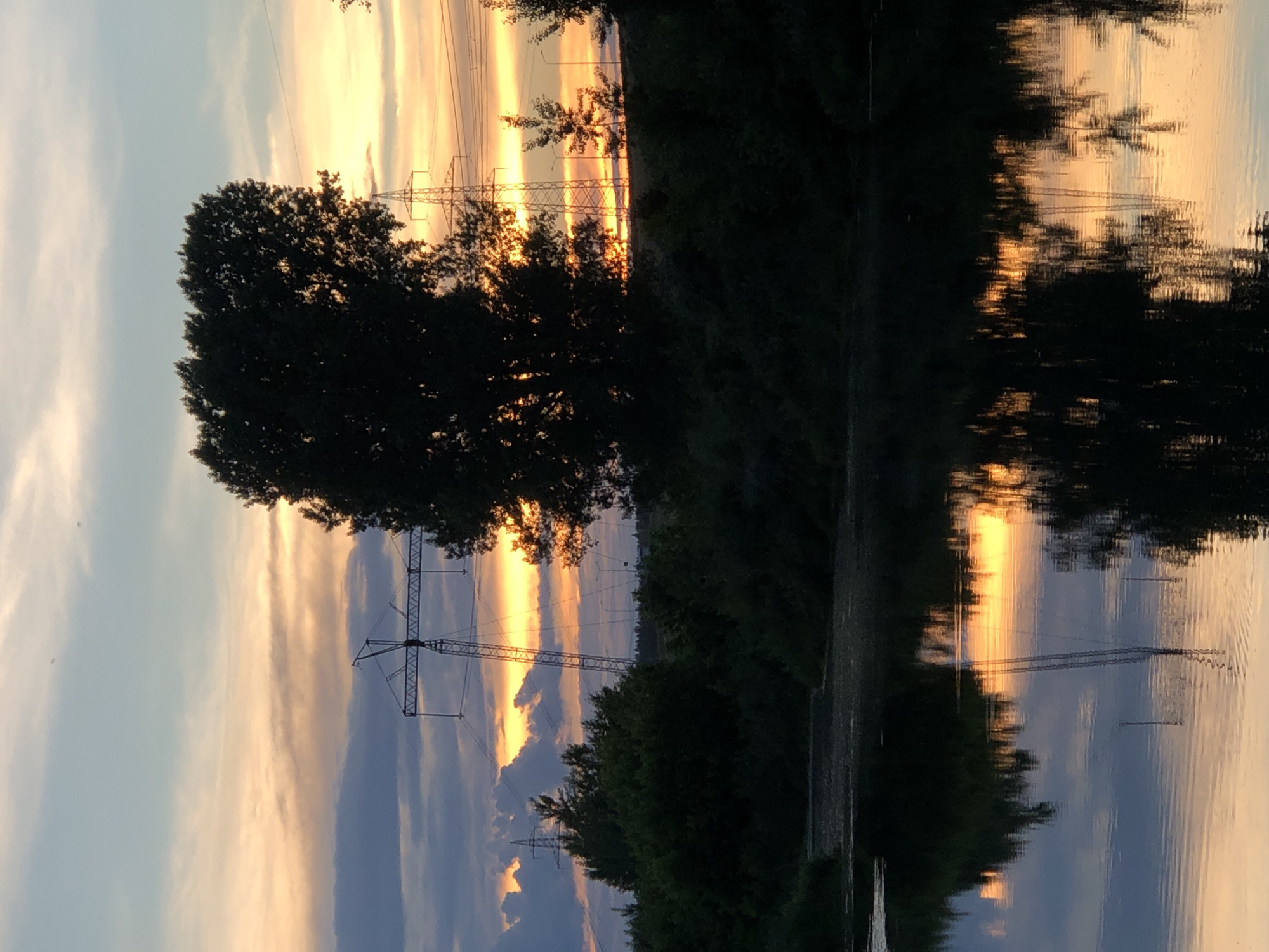 The image shows a sunset over the Kama River in Chaikovsky, Russia. The sky is filled with a mix of orange, yellow, and blue clouds. In the foreground, the dark silhouette of trees and a large transmission tower is reflected in the water. The water is calm, with ripples indicating a gentle breeze. The scene conveys a sense of tranquility and the beauty of nature.