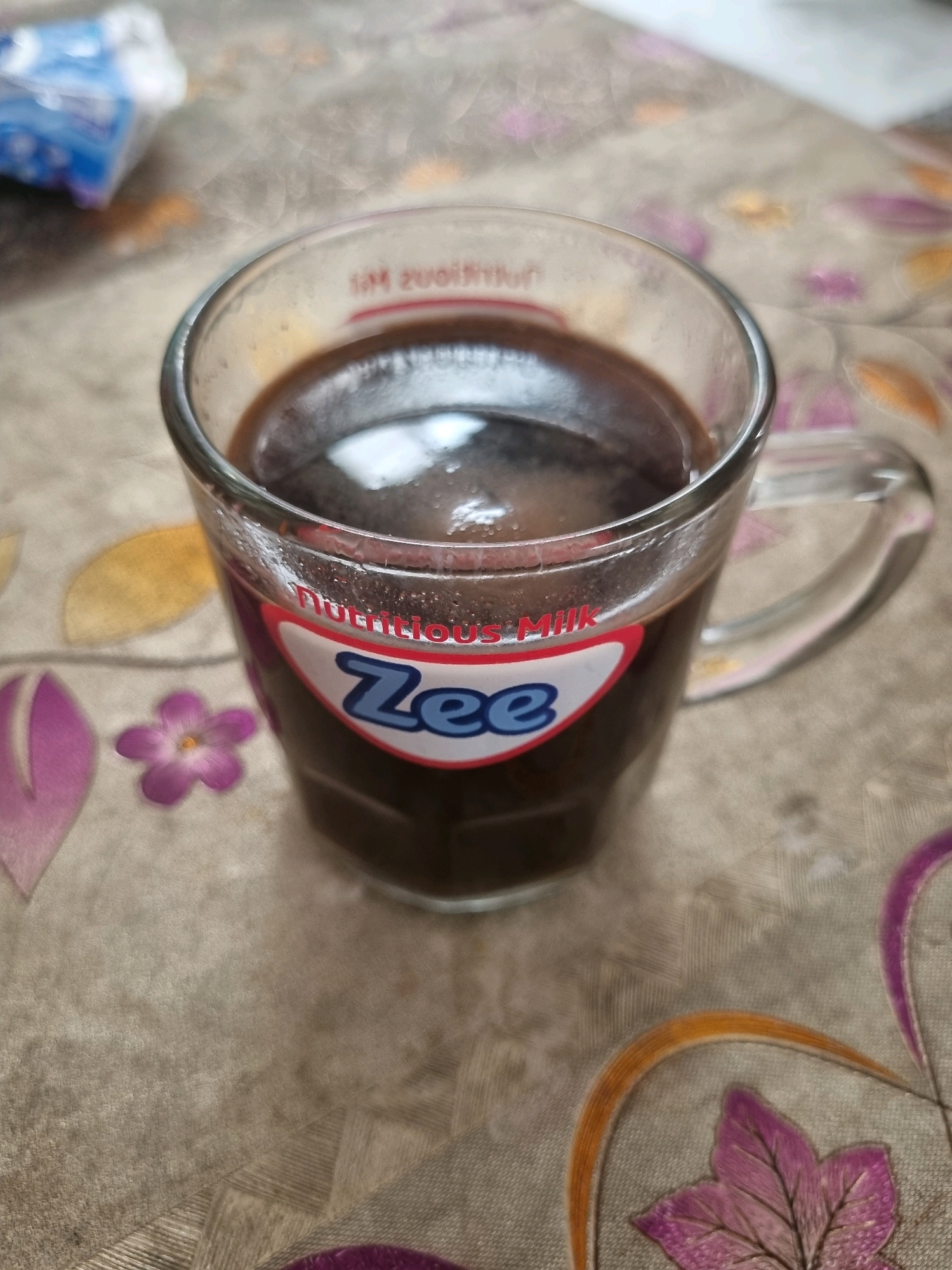 A clear glass mug containing a dark liquid, possibly coffee or hot chocolate, sits on a beige tablecloth with a floral pattern. The mug has a handle and the brand name "Zee" is visible on a red and white label. Some steam rises from the hot beverage. In the background, a blurry blue and white object, possibly a packet or box, is visible. The scene appears to be indoors, likely in a home environment.