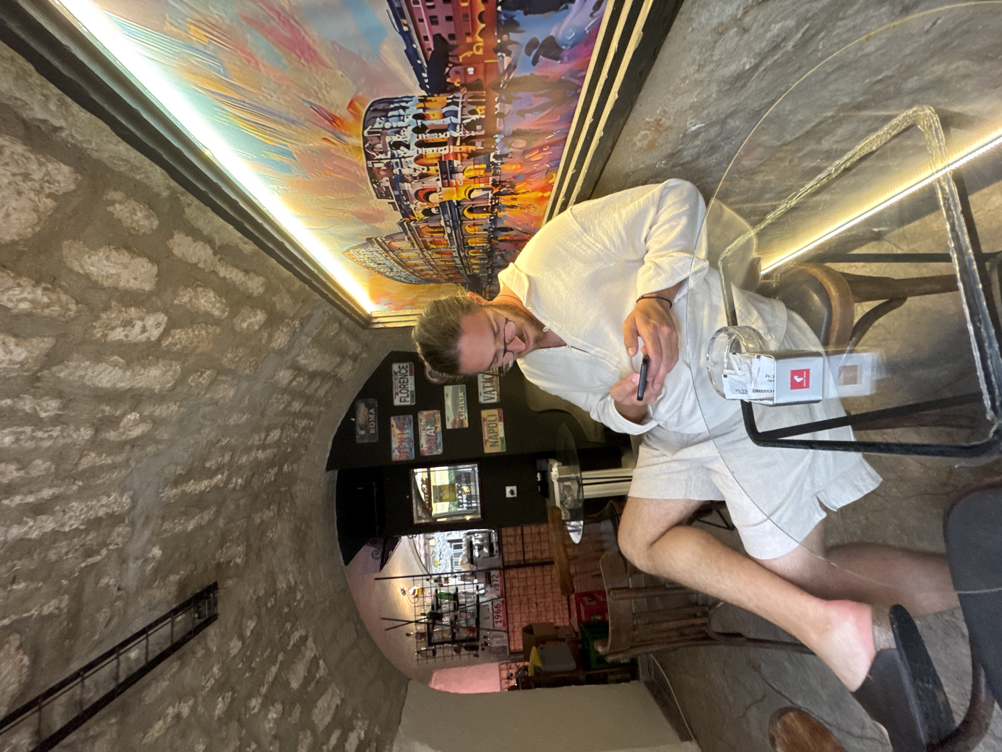 A man is seated at a glass-top table in a cafe with a stone arched ceiling and walls. He is wearing a light-colored, open-collared shirt and white shorts, and glasses. He is looking down at his phone, which he holds in his hands. To his right is a colorful painting of the Colosseum in Rome. There are several framed license plates on the wall behind him, with city names such as "ROMA", "FLORENCE", "CATANIA", "SICILIA", "NAPOLI", and "VATICAN". A bar area with shelves of bottles is visible in the background. The cafe appears to be located in Trebinje, Bosnia and Herzegovina.
