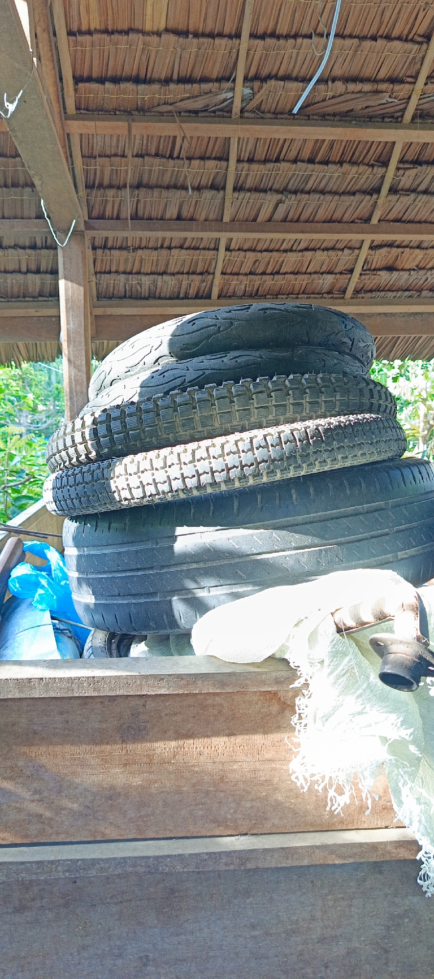 In Pashiebeutong, Indonesia, a stack of four tires rests on a wooden surface under a thatched roof. The tires vary in tread pattern and size, with the bottom tire appearing larger than the others. A blue plastic bag is visible behind the tires, along with some white fabric. The background shows greenery and sunlight filtering through the roof. The scene appears to be an outdoor storage area.