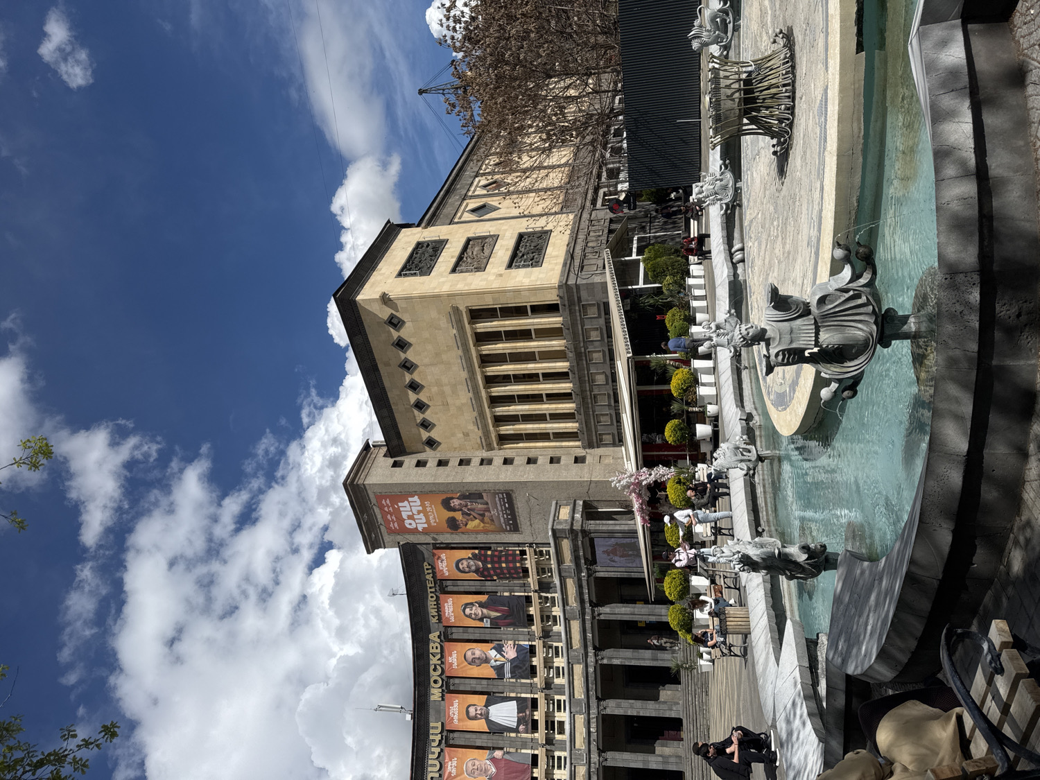 The image captures a lively public square in Yerevan, Armenia, bathed in bright daylight under a vivid blue sky with scattered white, fluffy clouds. The scene is dominated by a grand, multi-story building and an ornate fountain.
The building, located in the background and midground, features a light-colored facade with classical architectural elements, including columns, decorative friezes, and numerous windows. The lower section of this building is covered with large vertical banners and posters advertising films or events. Clearly visible text on these banners includes "MOCKBA КИНОТЕАТР" (Moscow Cinema) and "ՄՈՍԿՎԱ" (Moskva) in Armenian script, confirming the location as the Moscow Cinema. The posters display various actors or characters, with one prominent banner featuring Armenian text such as "ՈԼՈՒ."
In the foreground and midground on the right, a large, multi-tiered fountain is actively spouting water. Its design incorporates several ornate sculptures, possibly mythical figures or animals, from which water cascades into the basins. The water appears clear and a tranquil blue-green.
Numerous people are present in the square, engaging in various activities. Some individuals are seated at tables or benches around the fountain, possibly enjoying an outdoor cafe setting or simply relaxing. Others are seen walking or standing, adding to the bustling yet leisurely atmosphere of the public space. A person in a dark coat is distinctly visible in the lower right foreground.
Sparse tree branches are visible against the sky in the upper left, suggesting a change of season, while smaller green bushes and potted plants enhance the aesthetic around the fountain and building. The overall ambiance is calm and unhurried, reflecting a typical day in a central urban area.