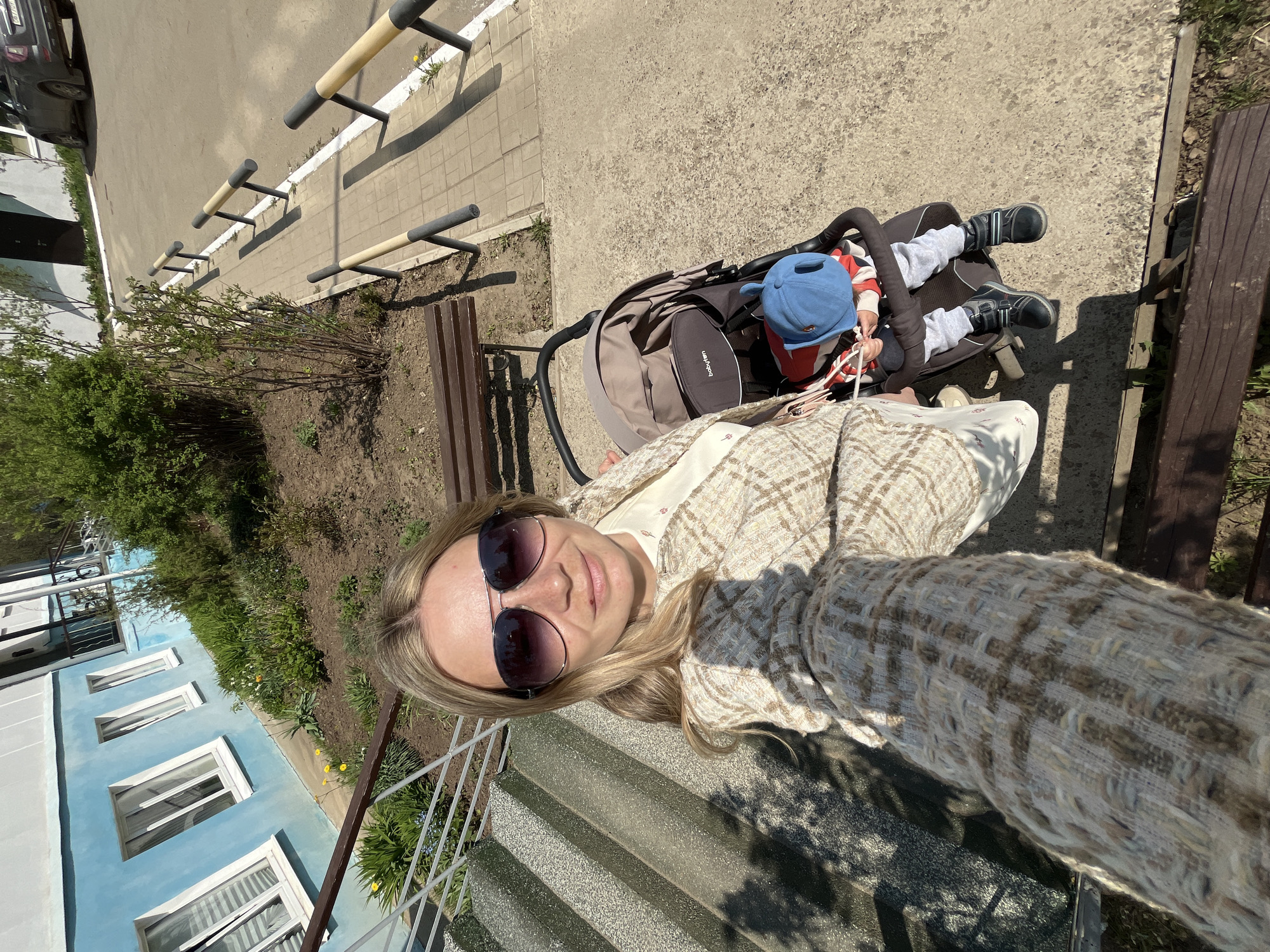 A woman in sunglasses and a patterned coat is taking a selfie on a set of stairs next to a metal railing. Behind her and to the right is a child in a stroller, wearing a blue hat, gray pants, and black shoes. A brown bench sits behind the stroller. In the background, there is a blue building with white-framed windows and a small garden area. To the right of the bench, there is a gray concrete walkway with metal barriers. Based on visual cues like architecture, vegetation, and concrete structures, the image is potentially located in the city of Chaikovsky, Russia.