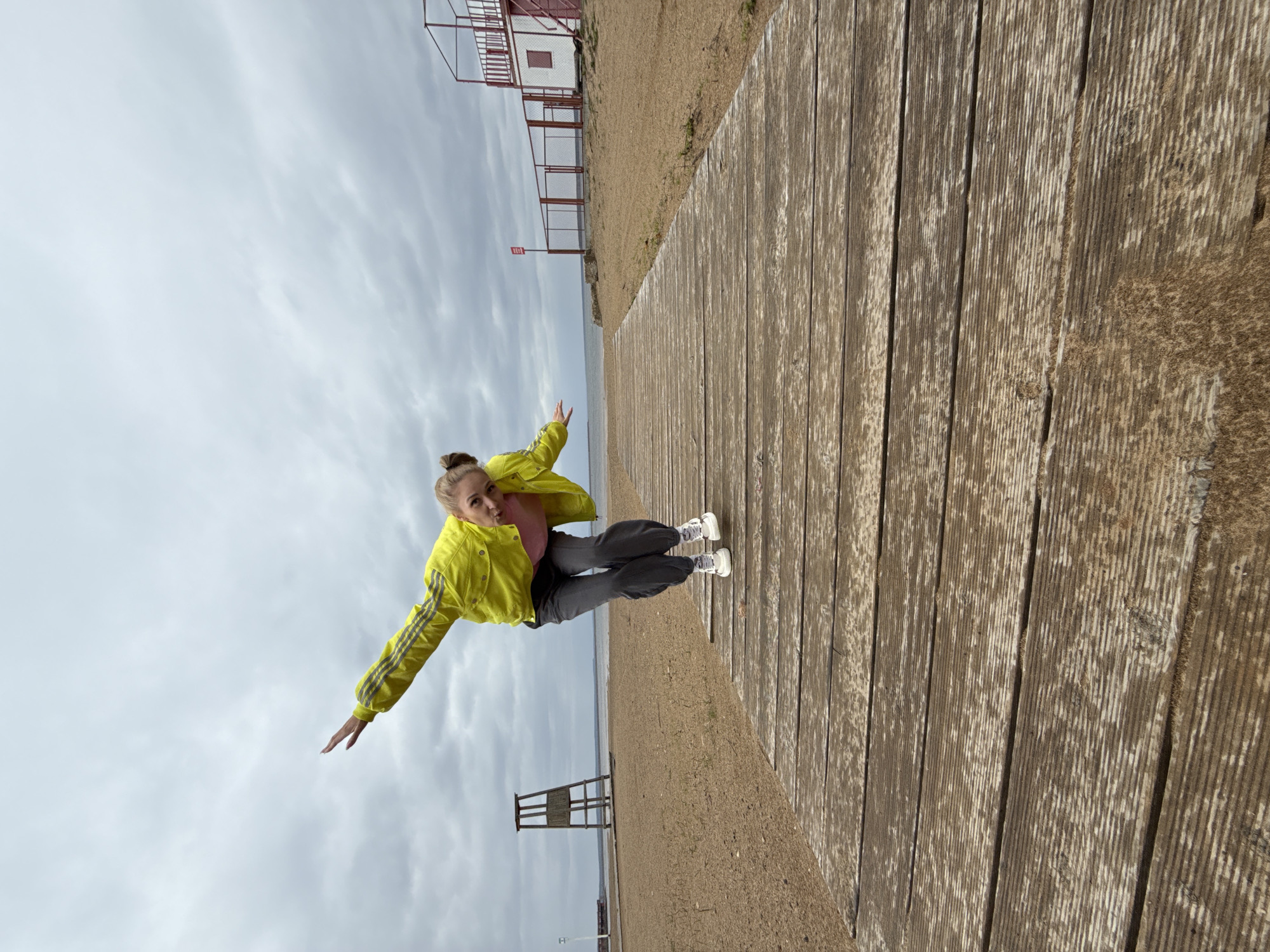 A young woman with light hair pulled into a bun is captured in a playful pose on a wooden boardwalk beside a sandy beach. She is wearing a bright yellow track jacket with dark stripes on the sleeves, a light pink top, dark grey pants, and white sneakers. Her arms are outstretched, and she has a playful expression with pursed lips, giving the impression of balancing or mimicking flight.
The scene features a long, weathered wooden boardwalk running diagonally into the background on the right side of the frame. To the left of the boardwalk is a stretch of sandy beach, which transitions into a calm body of water in the distance, likely a lake or large river. The sky above is overcast with numerous grey clouds, indicating a cloudy day.
In the mid-ground on the beach, a rustic wooden structure resembling a lifeguard stand or observation post is visible. Further in the background on the right, there is a more complex structure with red framing and white panels, possibly part of a building or a multi-level platform, accompanied by a fence. No discernible text is visible on any signs or objects. The overall setting suggests a recreational area by the water, consistent with a location like Chaykovsky, Russia.