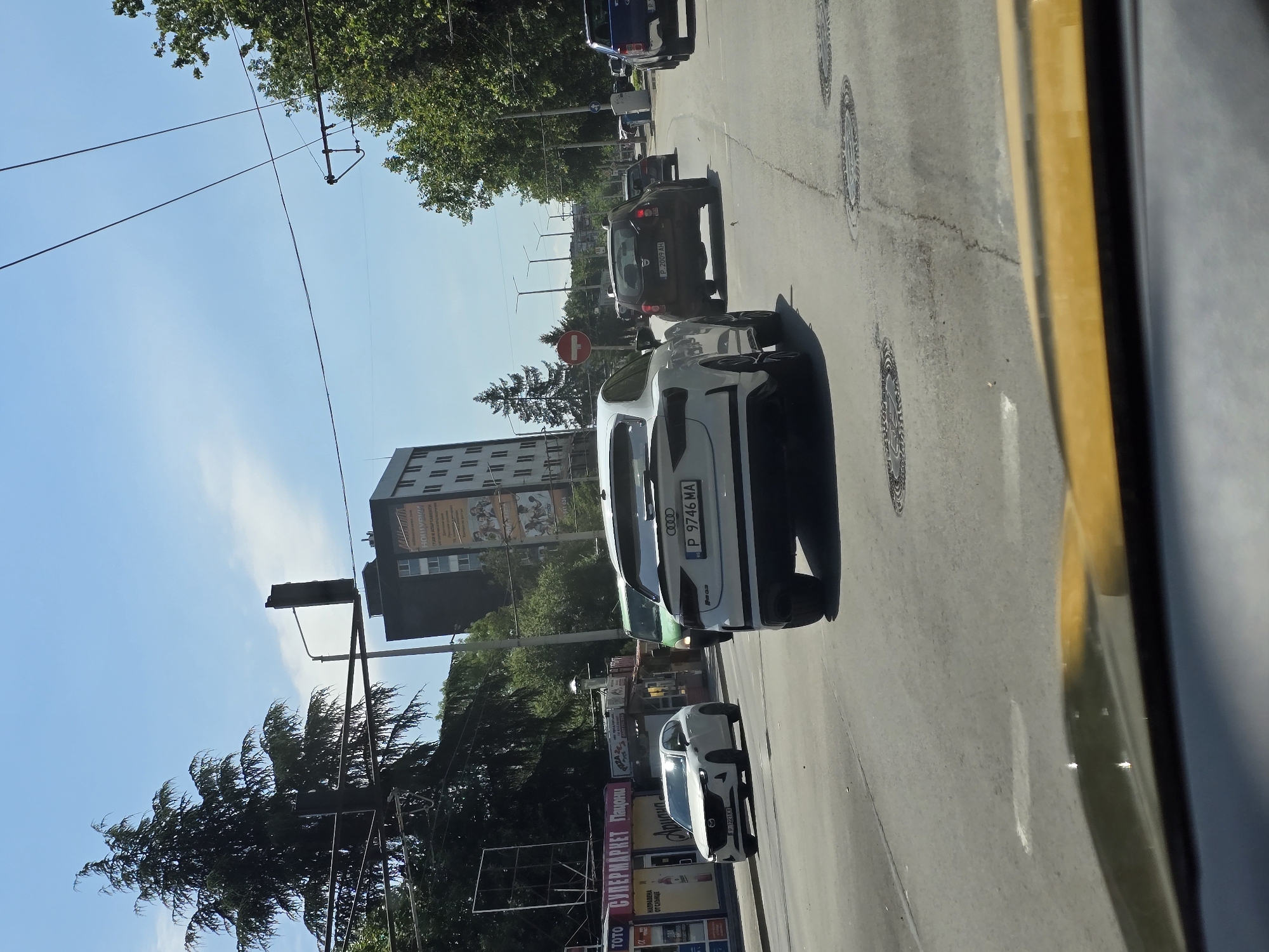 The image shows a street scene in Ruse, Bulgaria, on a sunny day. In the foreground, the dashboard of a yellow vehicle is visible. The road is paved with asphalt and marked with white lines. Several cars are driving on the road. A white Audi RS Q3 with Bulgarian license plate P 9746 MA is in the center of the image, facing away from the camera. To its left, a white Mazda CX-5 with license plate P 1221 KX is also visible. Further down the road, a dark-colored SUV, possibly a Dacia Duster with license plate P 2009 AH, is driving. A green van is also present.
On the left side of the road, there are shops with signs in Bulgarian, including "TOTO," "СУПЕРМАРКЕТ," and "ПАЦОНИ." A tall, dark-colored building with several floors is visible in the background, with advertisements on its facade. Trees line the right side of the road. Traffic lights and power lines are also present in the scene. The sky is blue with some clouds.
