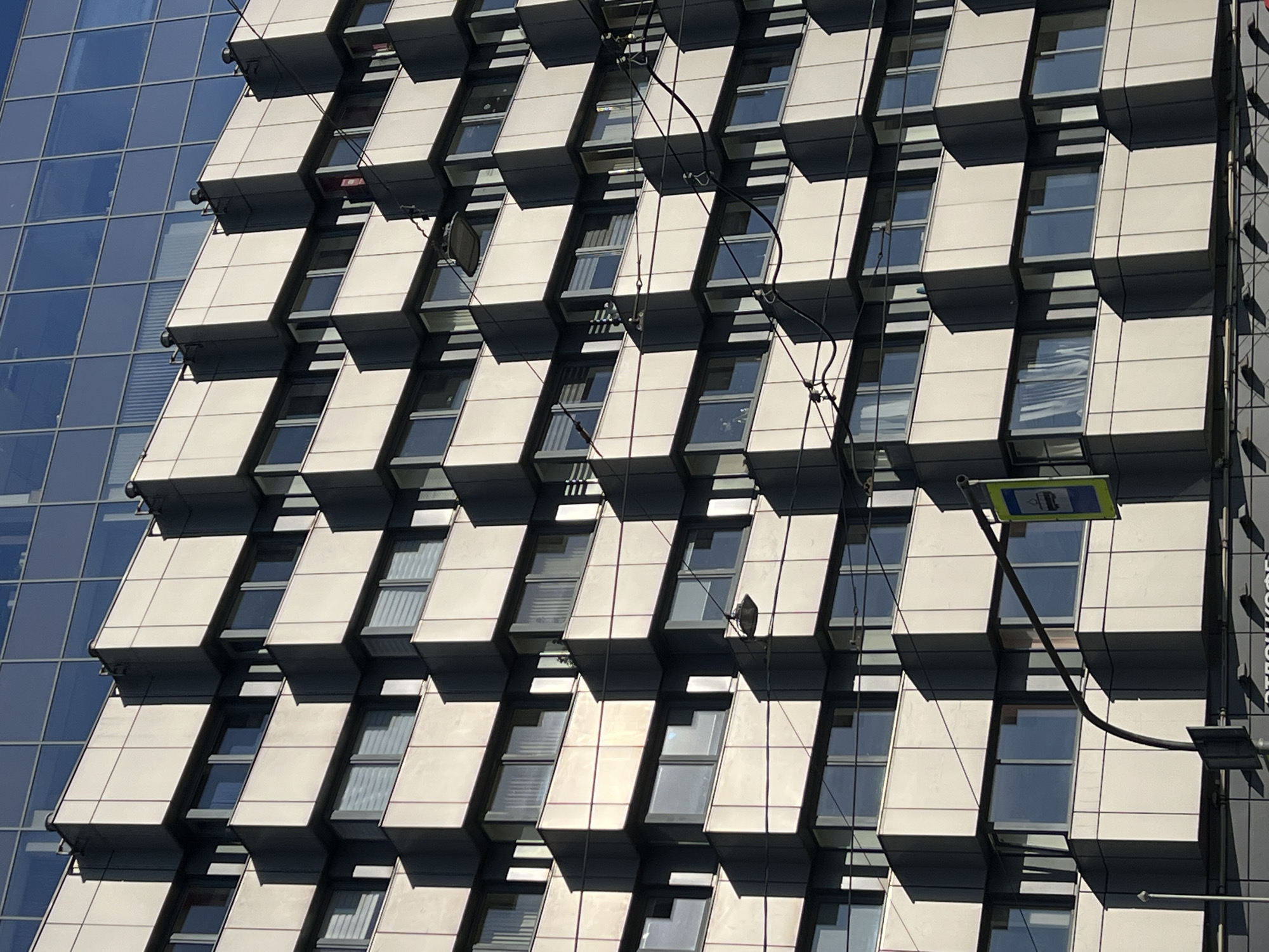 The image shows the facade of a modern building in Saint Petersburg, Russia. The building's facade is characterized by a pattern of rectangular blocks, some with windows, that protrude slightly from the building's surface, creating a three-dimensional effect. A smaller section of the building is visible to the left, featuring a more traditional glass curtain wall. Electrical wires and a sign are visible on the building's facade. The lighting indicates a sunny day, with shadows casting across the building.