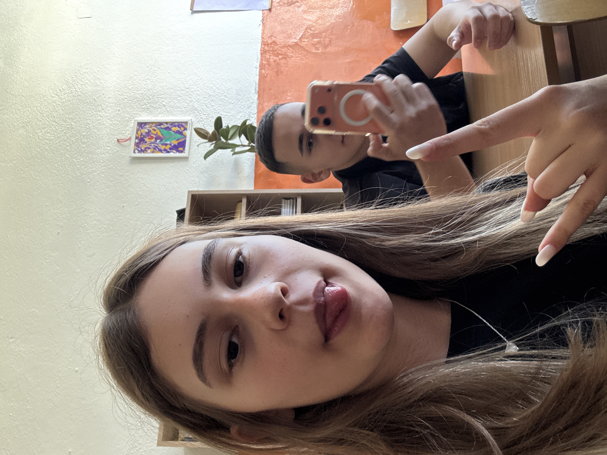 A selfie shows a young woman with long brown hair and light skin. She is wearing a black shirt and a delicate necklace with a triangular pendant. Her tongue is slightly sticking out. Behind her, a young man is visible, holding a smartphone with an orange case to take a photo. They appear to be sitting at a desk in a room with light yellow and orange walls. The background includes a small shelf, a green plant, and a framed picture on the wall. The location is identified as Panagyurishte, Bulgaria.
