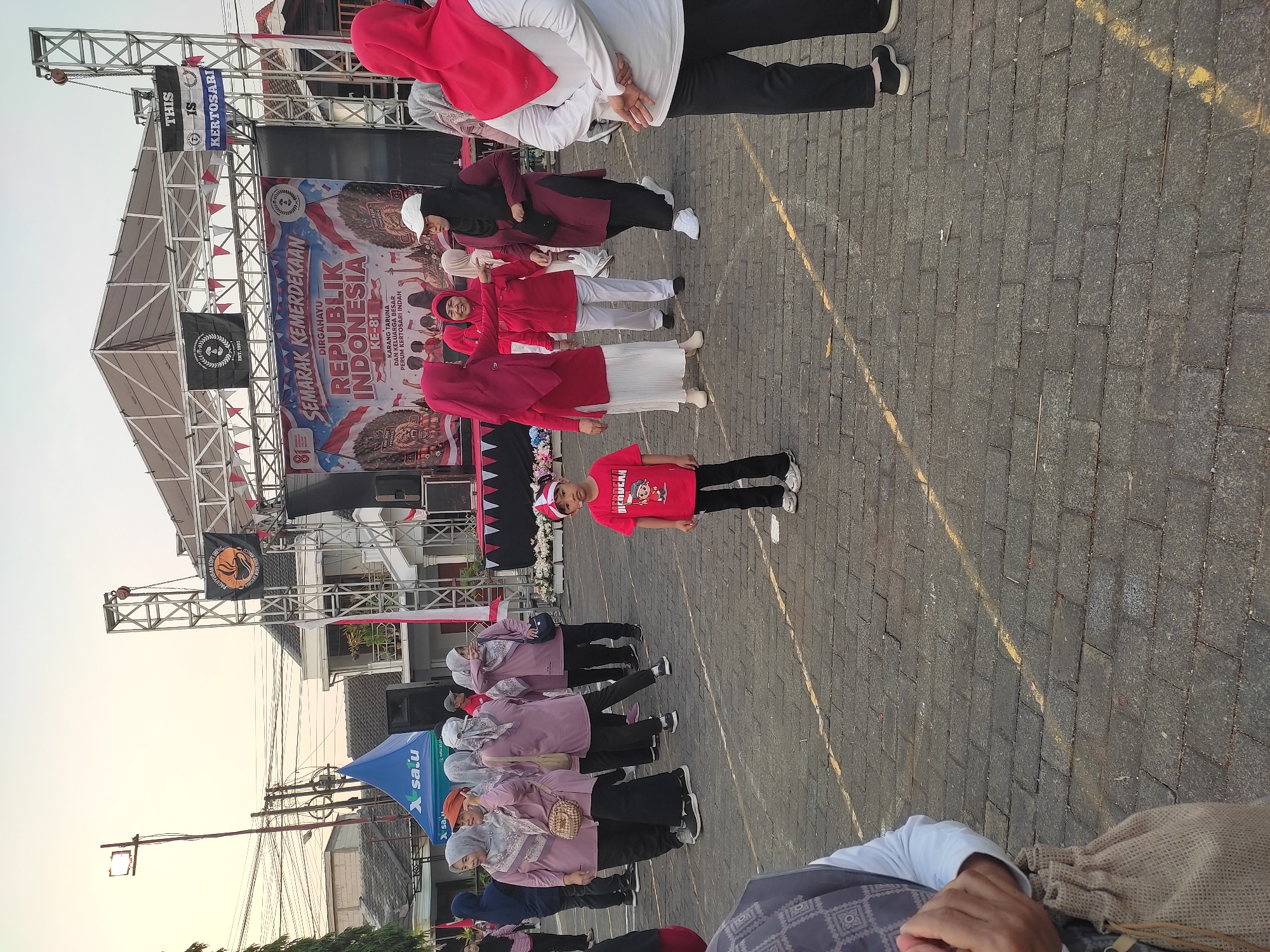 The image depicts an outdoor event likely celebrating Indonesian Independence Day, given the banner that reads "SEMARAK KEMERDEKAAN REPUBLIK INDONESIA". A stage is set up with a large banner and several flags. On the stage and in front of it, there are people dressed in various attire. In the foreground, a young boy in a red t-shirt and black pants stands facing the camera, wearing a red and white striped hat. A group of women, mostly wearing light purple tops and headscarves, are gathered to the left of the boy, some interacting with each other. To the right of the boy, more women are present, some in maroon outfits and headscarves, and one in a white shirt and black pants. The ground is paved with dark bricks, and yellow lines are visible, suggesting a marked area. The background shows buildings and trees, and the sky is a pale orange, indicating it might be early morning or late afternoon. The overall scene suggests a community gathering or celebration in an urban setting in Ponorogo, Indonesia.