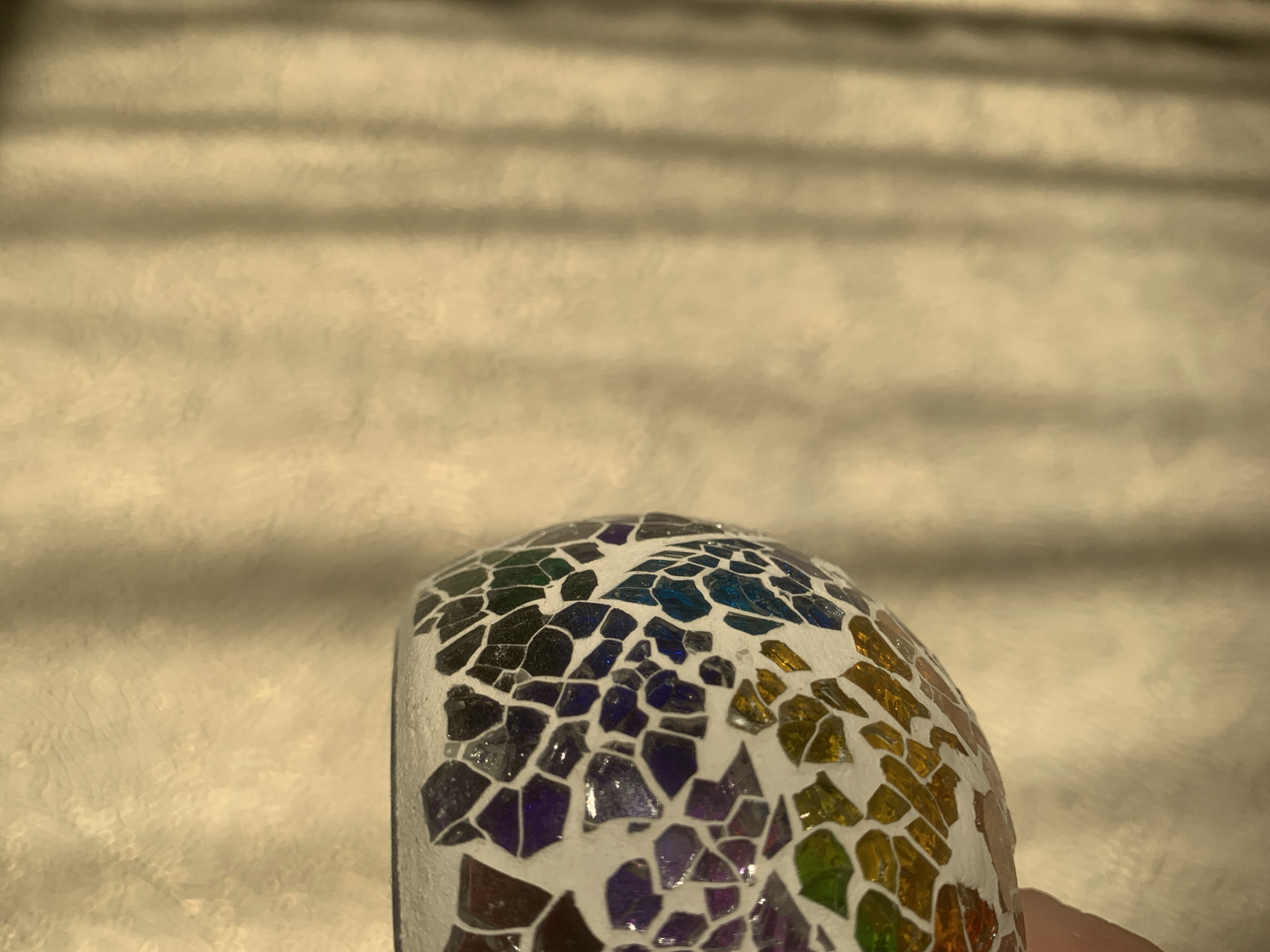 A close-up shows a small, rounded object covered in a mosaic of colorful glass or ceramic tiles. The mosaic features various colors, including black, blue, purple, orange, and green. The tiles are set in white grout. The background is a light, blurry surface. There is no indication of this being Izhevsk, Russia.