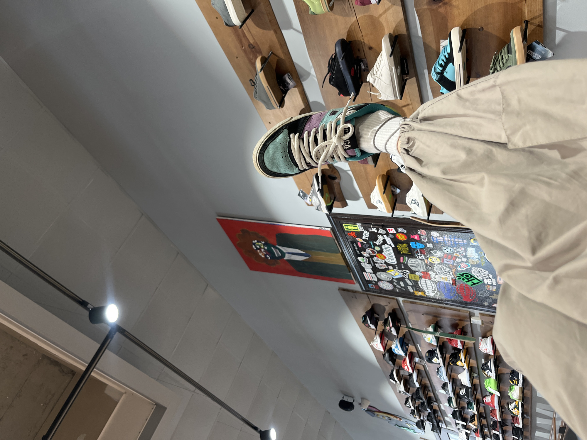 The image shows an indoor scene. The primary focus is on a person wearing a sneaker with a light green toe, purple midsection, and white laces. The person is wearing a white sock and beige pants. The backdrop features a wall display of various shoes on wooden planks. Also visible are two framed artworks, one with a red and black figure and another covered in stickers. The setting is likely a shoe store or boutique. The location is in Krasnoyarsk, Russia.