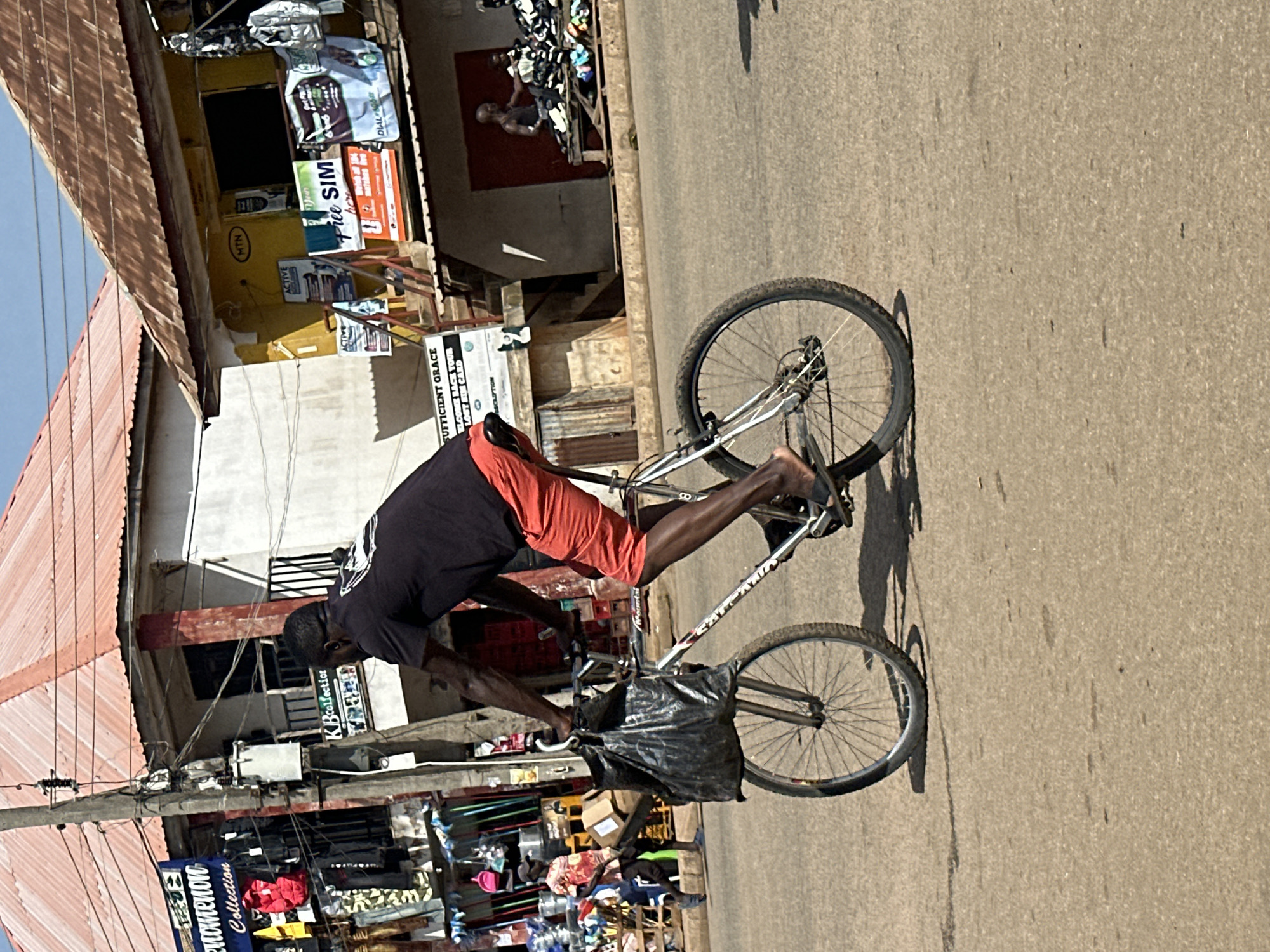 A Black man is riding a bicycle on a paved road in Bukuru, Nigeria. The man is wearing a black t-shirt and red shorts. His bicycle has a large black plastic bag hanging from the handlebars. The background shows a street scene with shops and buildings. One shop has a sign that says "MTN". Another sign advertises "FREE SIM". There are clothes hanging outside some of the shops. The sky is blue and the sun is shining.