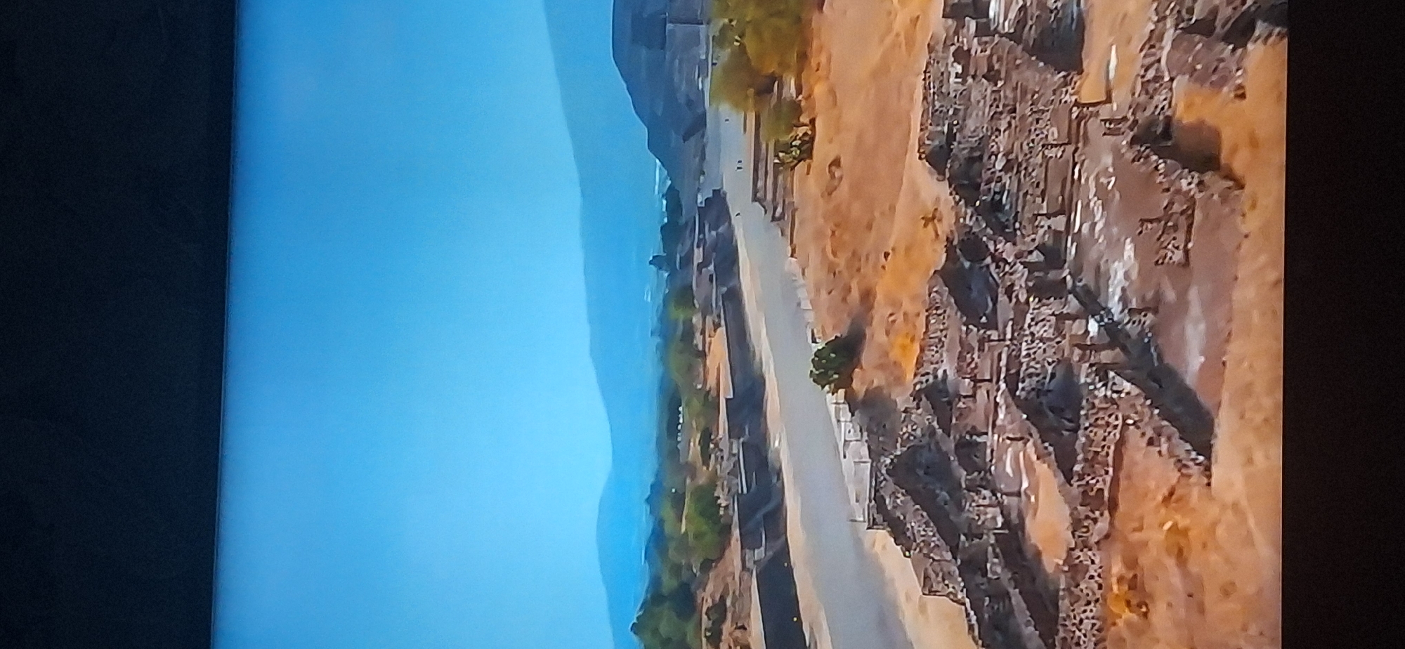 The image shows an aerial view of an archaeological site under a clear blue sky. In the foreground, there are ruins of ancient stone structures, characterized by walls and fragmented foundations. A paved pathway runs through the middle of the scene, separating the foreground ruins from more distant structures. Beyond the pathway, there are additional ruins and some scattered vegetation, including trees and cacti. In the background, a large, stepped pyramid structure is visible, hinting at a significant historical settlement. Further in the distance, rolling hills and mountains form the horizon line. The overall environment is arid and sunny, with evidence of ancient construction. This site does not appear to be located in Bila Tserkva, Ukraine.