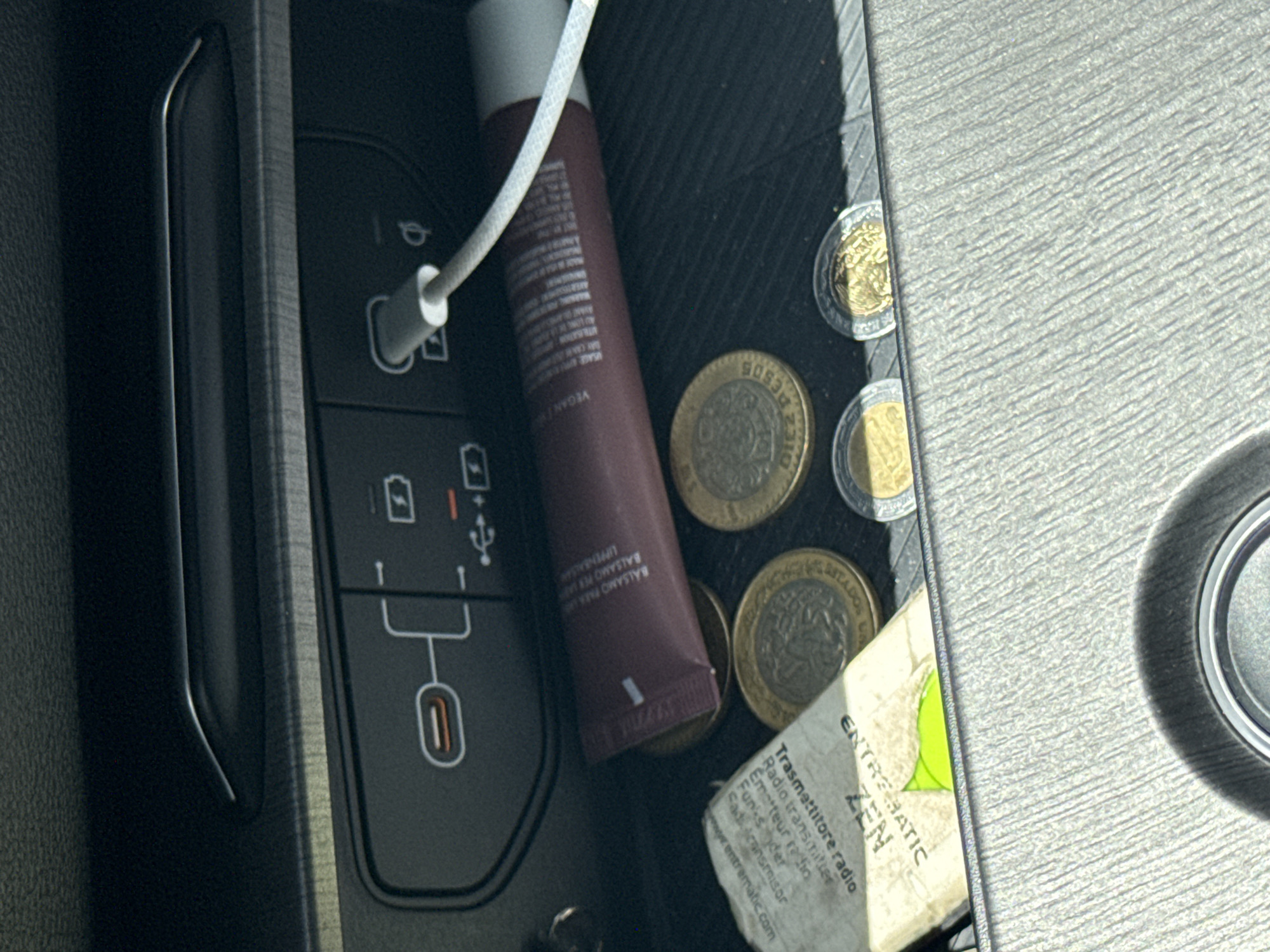 The image shows the center console of a car, likely a modern vehicle due to the USB-C and USB-A ports visible. A white charging cable is plugged into the USB-C port. Next to the charging ports is a tube of lip balm with writing including "VEGAN." Several Mexican coins are scattered around the lip balm and a small piece of paper. The paper is partially visible and contains the words "ENTREMATIC ZEN" and "Trasmettitore radio" (Radio Transmitter). The website address "www.entrematic.com" is also on the paper. The environment appears to be inside a car, possibly in Puebla, Mexico, based on the currency.