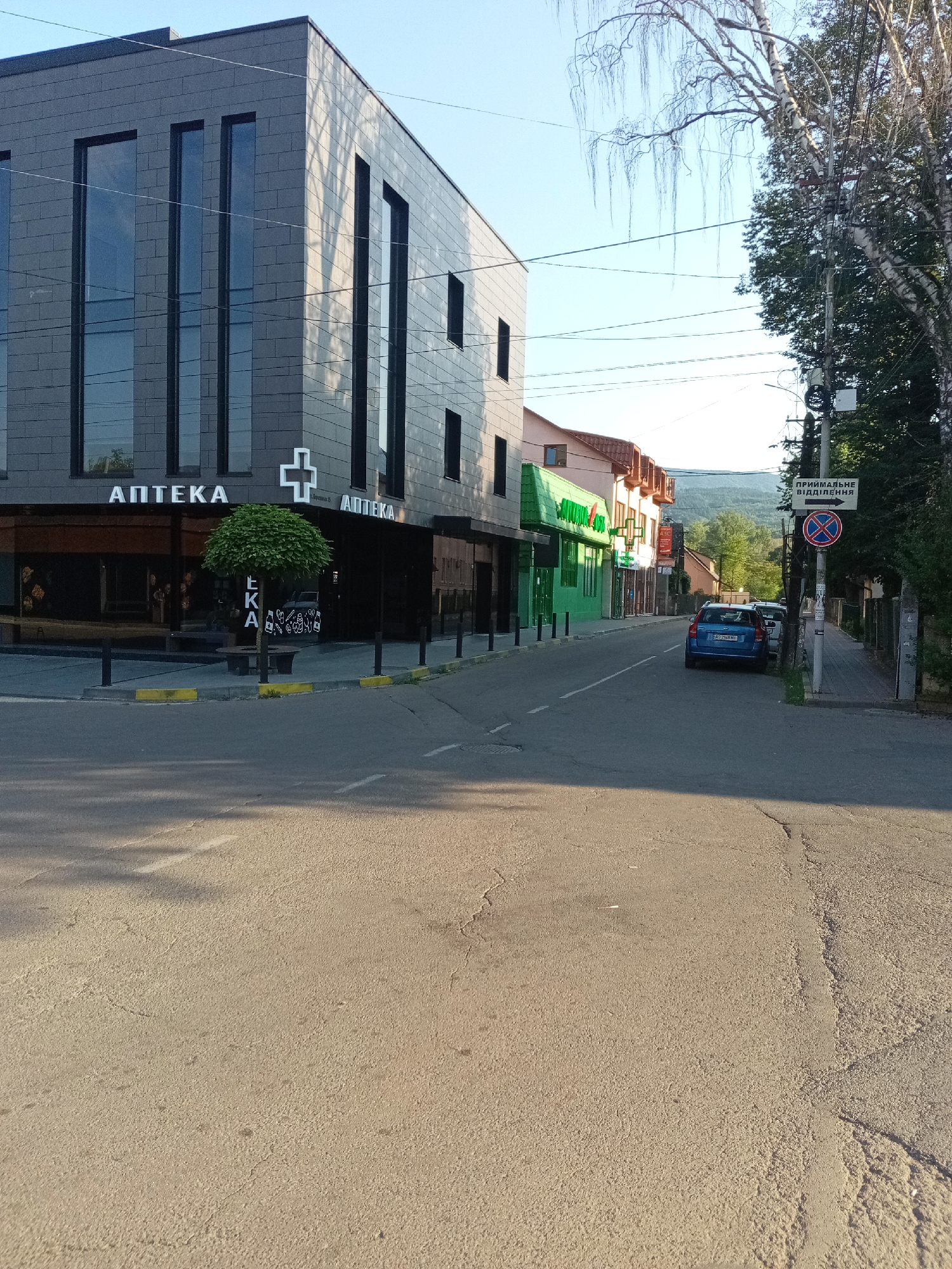 The image depicts a street scene in Tiachiv, Ukraine, on a clear day with sunlight casting shadows. On the left, a modern, gray building with dark framed windows dominates the foreground. The building has signage in Cyrillic script, including "АПTEKA" (Apteka, meaning pharmacy) displayed multiple times, along with a prominent white cross symbol, a common emblem for pharmacies. A small, round-top tree with dense foliage is planted in front of the building, adjacent to a bench.
The street is paved and appears to be a public road with a pedestrian sidewalk visible on the right. Several cars are parked along the right side of the street, including a blue sedan and a white car. A street sign with Cyrillic text and an arrow points to the right, indicating "ПРИЙМАЛЬНЕ ВІДДІЛЕННЯ" (Priimalne viddilennya, meaning reception or intake department), accompanied by a traffic sign prohibiting stopping or parking.
Further down the street, other buildings are visible, including one with a bright green facade and signs that are partially obscured. The background shows a landscape with trees and hills, suggesting a semi-urban or suburban setting within Tiachiv. Overhead, power lines crisscross the sky, and a birch tree with distinctive white and black bark stands on the right side of the street. The overall impression is a typical day in a Ukrainian town.