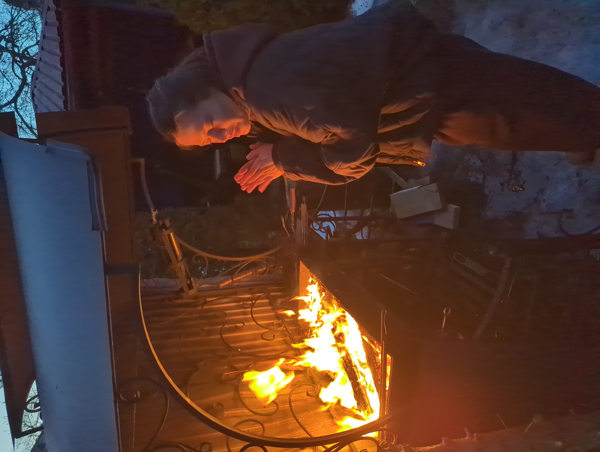 An outdoor scene is depicted at dusk or night in a backyard setting in Khimki, Russia. A lone female individual, wearing a dark, padded jacket and dark trousers, stands positioned to the right of a large, ornate metal brazier. Her hands are clasped together, palms facing the brazier, indicating she is warming them; her face, illuminated by the firelight, shows closed or squinted eyes. The brazier contains an active wood fire with multiple burning logs emitting prominent yellow and orange flames and a significant glow. Above the brazier, a dark metal canopy or hood with decorative scrollwork is visible. The ground surface beneath the subject and around the brazier exhibits patches of snow or ice. In the background, bare tree branches are silhouetted against a dark blue sky, alongside a portion of a building with a dark, corrugated metal roof.