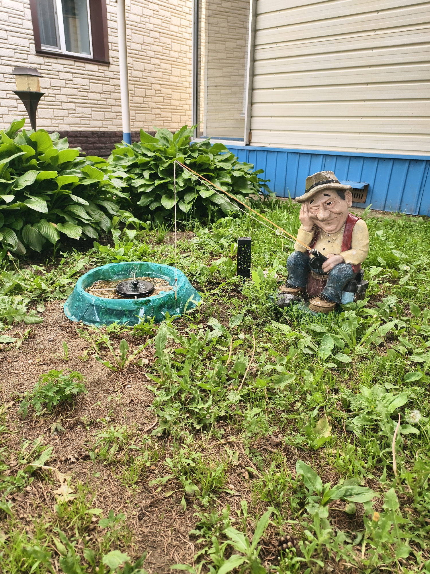This media file depicts an outdoor garden scene in Belogorsk, Russia. The primary subjects are a garden gnome figurine and a small, active fountain. The gnome, made of resin, is designed to resemble an elderly man wearing a hat, a vest over a light-colored shirt, and jeans. He is seated, holding a fishing rod that extends towards the fountain, suggesting he is "fishing." His expression is slightly weary or contemplative.
The fountain is a shallow, teal-colored basin with water spraying upwards from a central mechanism. The fountain is surrounded by lush green foliage, likely hostas, and a patch of lawn that is interspersed with weeds and dry earth. In the background, a house with beige, textured siding and a white window is visible. A blue trim runs along the base of the house.
The scene appears to be set during daylight hours, with natural lighting illuminating the area. The weather seems temperate, suitable for an outdoor setting. The overall atmosphere is relaxed and perhaps a touch whimsical, owing to the garden gnome and its fishing activity. There is no visible text in the image.