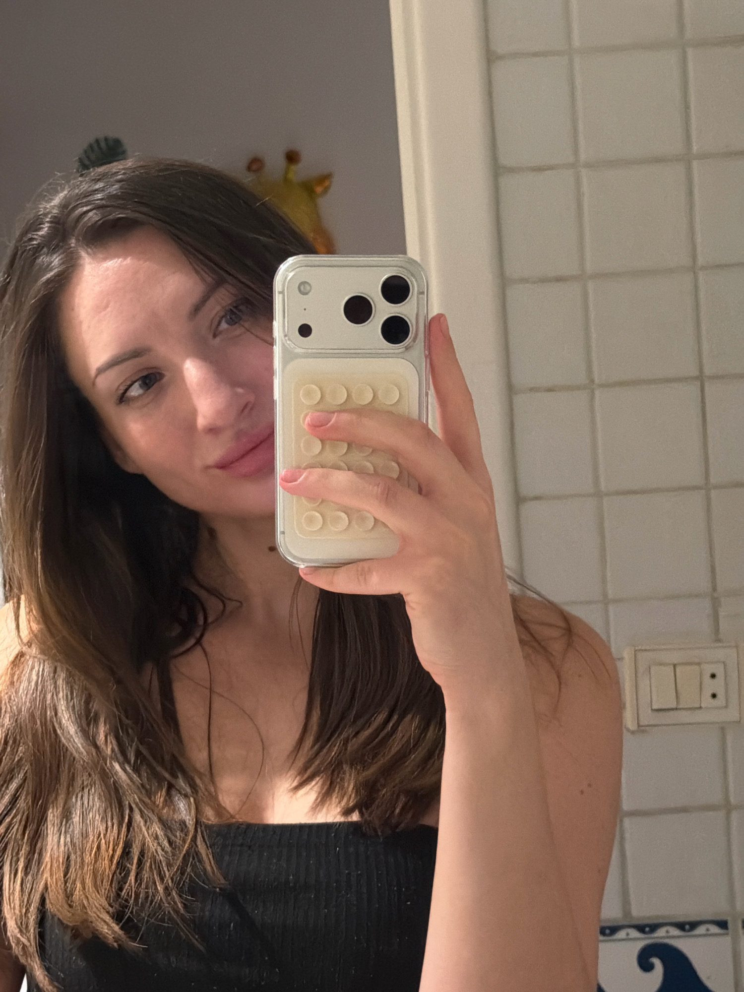 A woman is taking a mirror selfie in a tiled bathroom. She has long, dark brown hair and is wearing a black top. She is holding her phone, which has a white case with a dimpled texture, in front of her face. In the reflection, a yellow giraffe decoration is visible on the wall behind her. The bathroom tiles are white and square. A light switch with two toggles is mounted on the wall to the right. A blue and white patterned border is visible at the bottom of the tiled wall. The overall scene suggests a personal, private space within a home or hotel room.