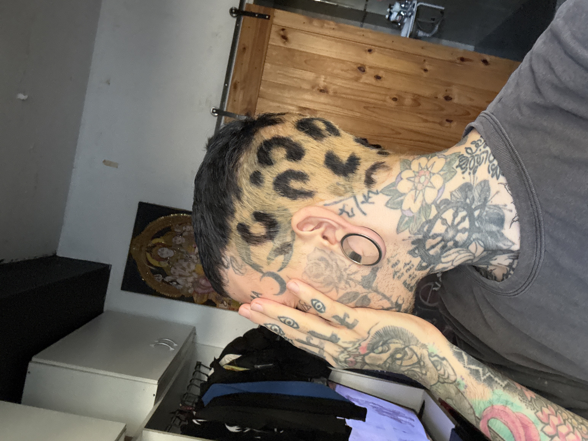 A person with extensive tattoos on their neck, face, and arms is positioned indoors with a hand covering part of their face. The person has a leopard print design shaved into their hair. They are wearing a grey t-shirt. A white storage cabinet is visible in the lower left. Clothes are piled on a rack below. A framed circular decoration hangs on the wall behind the person. A wooden sliding door is in the background. It is not possible to determine the specific location in Buenos Aires, Argentina from this image.