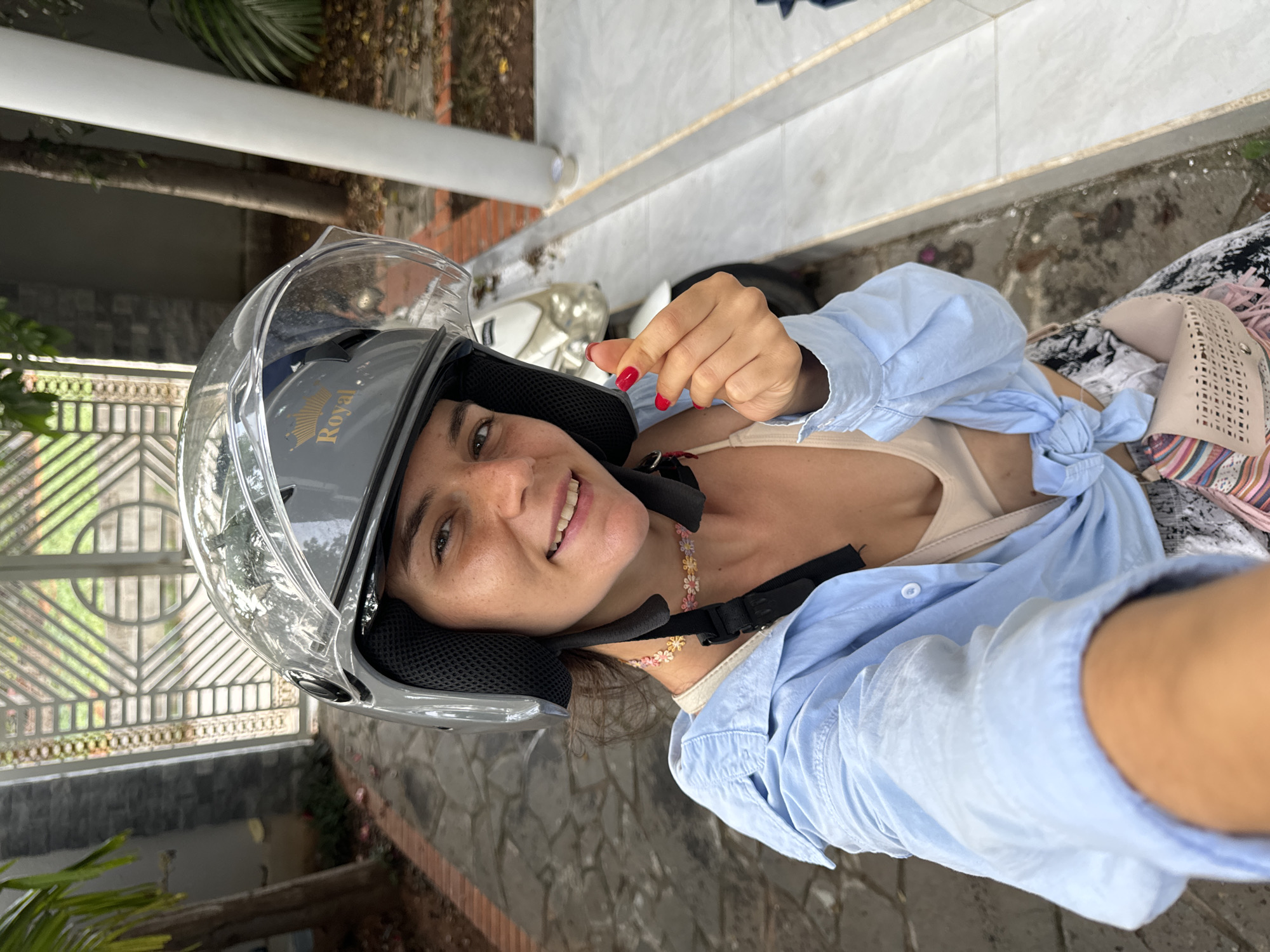 This is a vertical, eye-level, close-up selfie of a young woman wearing a silver motorcycle helmet with a clear visor. She is smiling broadly at the camera and making a hand gesture with her right hand, which has bright red nail polish. She is wearing a light blue, unbuttoned shirt tied at the waist, revealing a beige tank top underneath. A colorful floral necklace is visible around her neck. The background shows a stone-tiled ground and some greenery, including a palm frond. A white motorcycle is partially visible behind her. The overall impression is casual and cheerful, likely taken outdoors on a sunny day, possibly in Mũi Né, Vietnam. The helmet has "Royal" written on it in gold lettering.