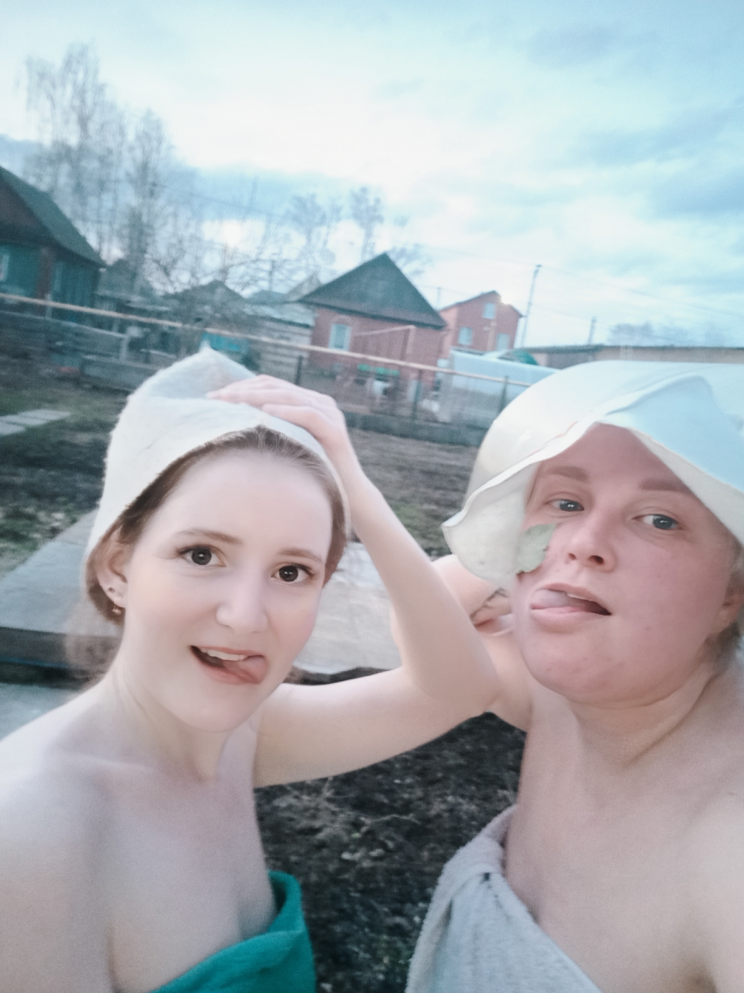 Two women are posing for a selfie in what appears to be an outdoor setting. Both are wearing towels and head coverings, possibly bath hats. One woman is wearing a green towel, and the other is wearing a white towel. The woman on the left is sticking out her tongue. The woman on the right is also sticking out her tongue and has a leaf or a similar object on her face.
In the background, there are houses and trees. The sky is overcast. According to metadata, this image was captured in Chaikovsky, Russia.