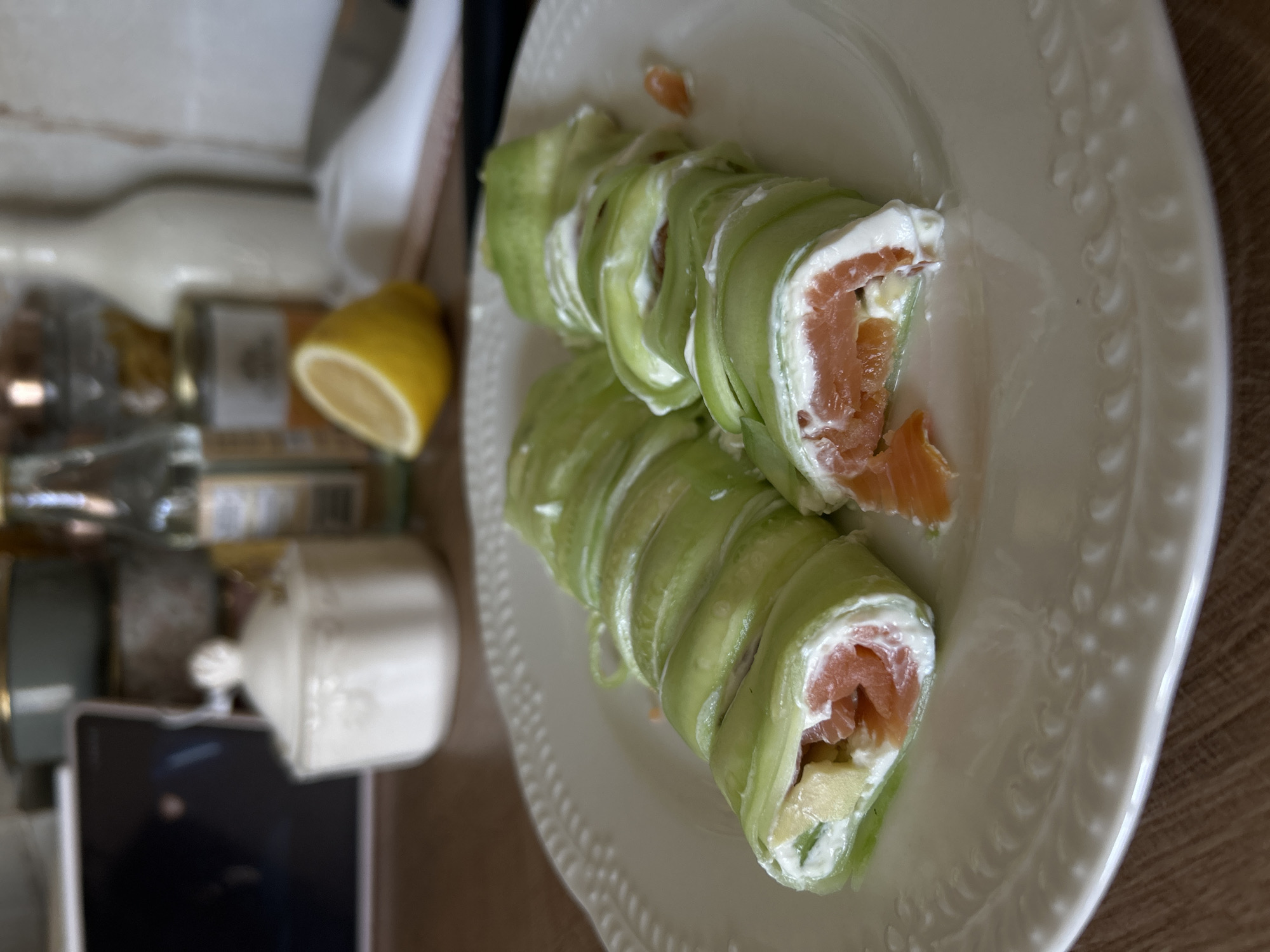 This media file features a close-up, eye-level shot of a white, embossed plate holding two cucumber rolls filled with smoked salmon, cream cheese, and what appears to be avocado or yellow bell pepper. The rolls are presented on the right side of the frame, with the plate occupying most of the space. The background is blurred but suggests a kitchen or dining area with wooden surfaces.
To the left of the plate, a halved lemon is visible, along with several bottles and jars, hinting at ingredients or condiments. The lighting appears to be natural, coming from above and slightly to the left, casting soft shadows. The overall scene is still and focused on the food. There is no visible text in the image. The setting is likely a home kitchen in Plovdiv, Bulgaria, given the provided location context. The meal appears to be prepared for consumption.