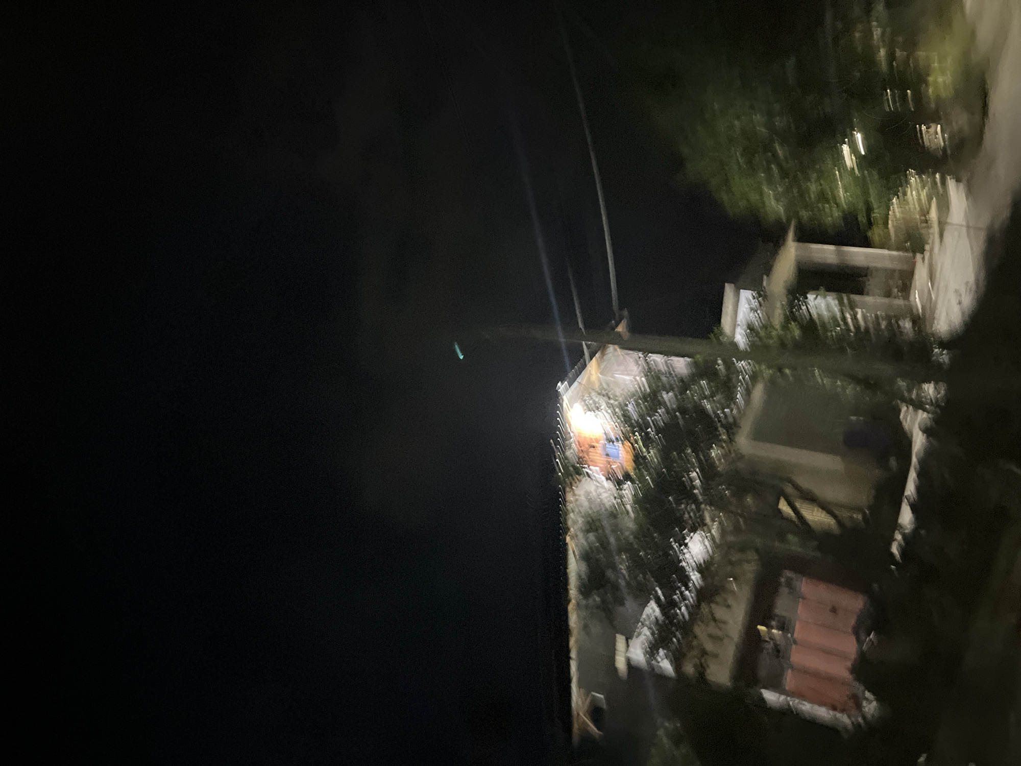 The image, taken at night in Menanggini, Indonesia, captures a slightly blurred view of a building and its surroundings. The scene is dimly lit, with some lights emanating from the building itself, suggesting interior illumination or outdoor lighting. The building appears to be multi-story with visible structural elements like beams and walls. There's a section of the building with a reddish, possibly tiled, facade.
Surrounding the building are trees and foliage, also partially obscured by the darkness and motion blur. Power lines are visible in the frame, stretching across the dark sky. The ground area near the building seems to be a pathway or a yard.
The overall impression is one of a residential or commercial area at rest during the night. The blurriness suggests the photo might have been taken from a moving vehicle or with a shaky hand. There are no people clearly visible, and the focus is on the architectural elements and the natural surroundings under the cover of darkness. The time of day is definitively night due to the deep shadows and the reliance on artificial light sources. No text is discernible in the image.